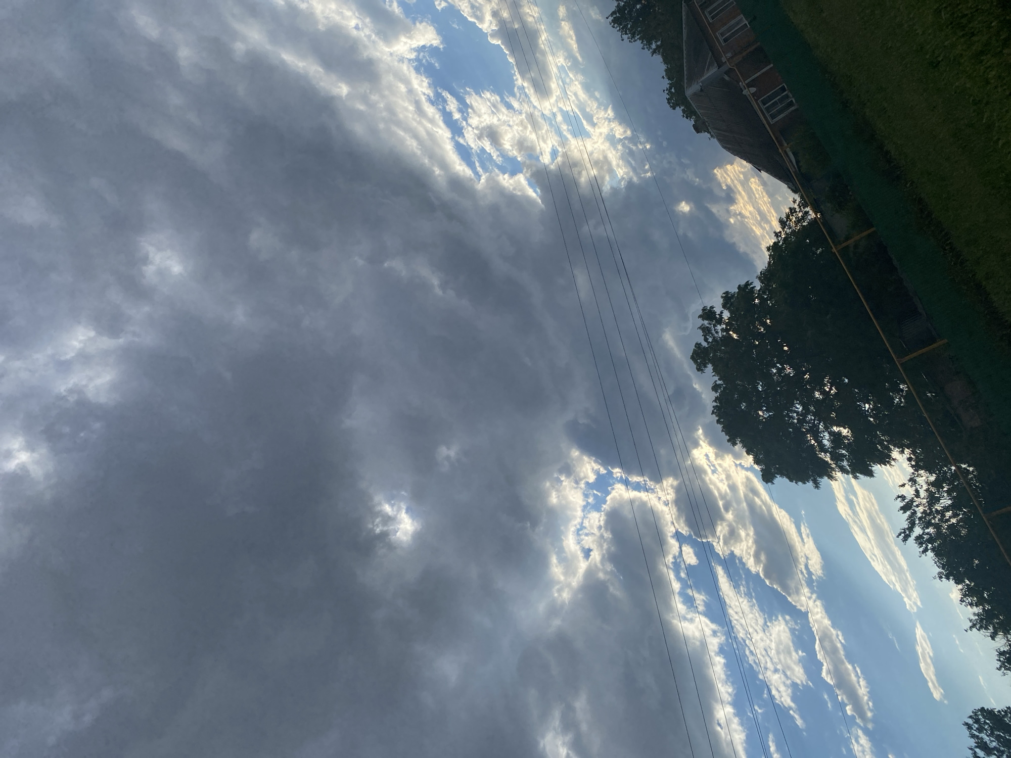 The image shows a dramatic sky filled with dark, heavy clouds interspersed with patches of bright blue sky illuminated by sunlight. Rays of light pierce through the clouds in some areas. The perspective is from ground level, looking upwards and slightly to the right. In the foreground, there is a grassy slope with a yellow-topped fence. To the right, a small, rustic house with a corrugated roof is visible, partially obscured by trees. A large, dark tree is prominent in the middle ground. Power lines stretch horizontally across the upper portion of the image. The scene appears to be in an outdoor, possibly rural or suburban, setting. There are no people visible in the image. The environment suggests a possibly overcast day with the sun breaking through.