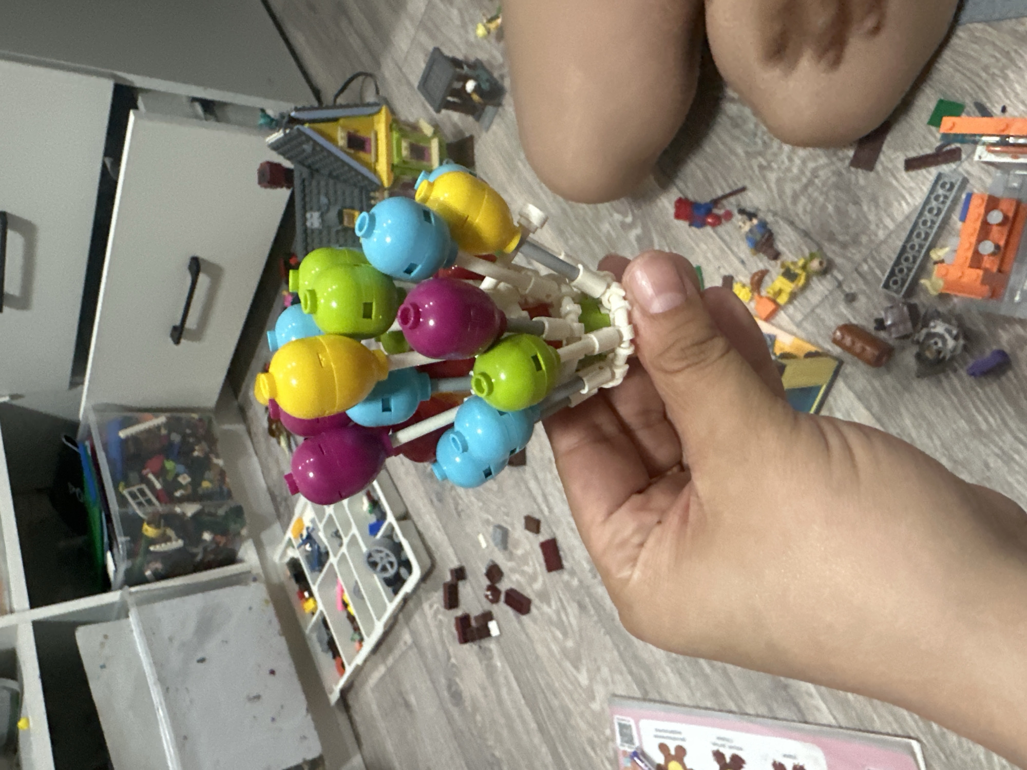A first-person view shows a hand holding a partially-assembled Lego Duplo creation resembling a cluster of colorful balloons or baubles. The object consists of green, yellow, blue, and purple rounded Lego pieces connected by white connectors. Scattered around the hand are various Lego pieces of different colors and sizes, including minifigures and building blocks. The background features a grey, wood-patterned floor, white storage shelves filled with Lego sets, and a partially visible pair of knees. A pink piece of paper with cartoon bears is also visible.
