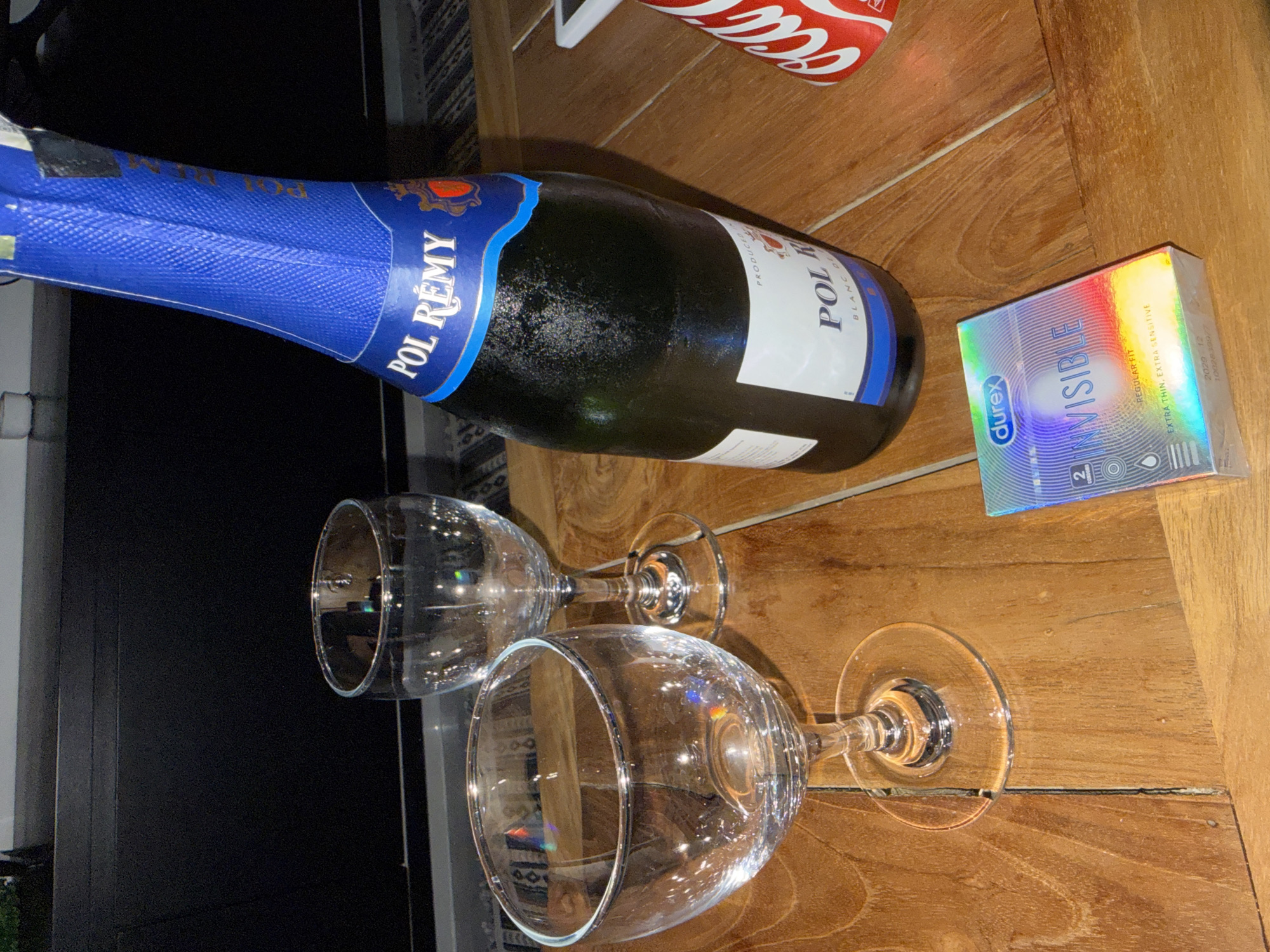 On a wooden table, likely indoors, a partially visible Coca-Cola can sits next to a bottle of Pol Remy sparkling wine. Two empty wine glasses are positioned below the bottle. A package of Durex Invisible condoms is placed to the right of the bottle. The scene suggests an intimate setting. The location in North Kuta, Indonesia is not definitively indicated within the image; this is inferred from external knowledge.