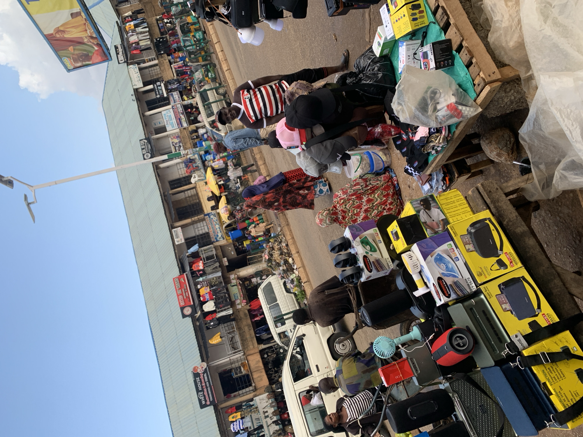 The image depicts a bustling street market scene in Bukuru, Nigeria. In the foreground, a variety of electronic goods, including speakers, fans, and irons, are displayed on wooden stalls and the ground. Several people are engaged in activities related to the market. A man in a striped shirt and black pants stands near a pile of caps, smiling at the camera. Another man is seated on a cart laden with more electronics. A woman in a black and white striped top is also seated nearby. In the background, a white van is parked on the street, and other vehicles, including what appear to be three-wheeled taxis, are visible further down the road. Shops with various signage, such as "WHITE HOUSE Cosmetics & Beauty" and "SNAP FIT WEARS," line the buildings along the street. The overall environment suggests a vibrant commercial area with a mix of retail shops and street vendors.