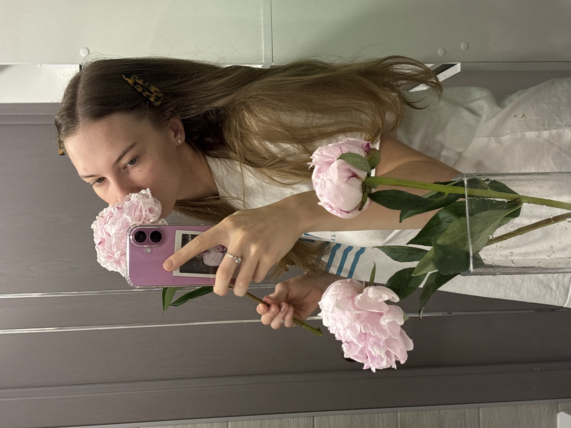 A young woman with long brown hair, wearing a white t-shirt and a ring on her left hand, stands in front of a mirror, taking a selfie. She holds a bouquet of pink peonies in her left hand, bringing one of the flowers to her nose to smell it. Her right hand holds a pink smartphone, with a picture of what appears to be a child or a small person visible on the back of the phone case. Two tortoiseshell-patterned hair clips are placed in her hair. The background appears to be a bathroom, with a gray tiled wall on the left and a lighter gray, possibly painted, wall on the right. A clear glass vase containing the peonies and water is visible in the foreground. The lighting suggests an indoor setting, likely a domestic bathroom. There is no identifiable information to suggest the location is in the city of Perm, Russia.