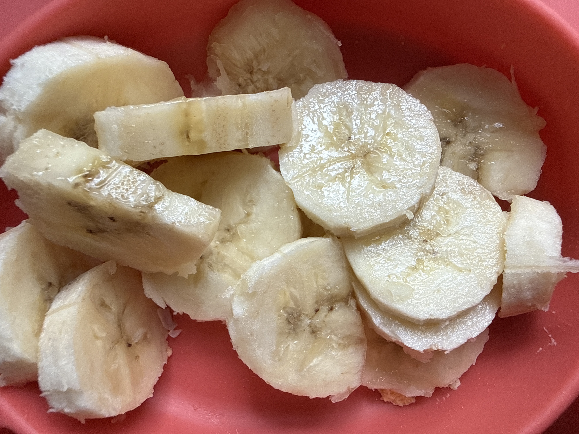 A close-up, overhead shot displays a collection of fresh banana slices resting in a reddish-orange bowl. The bananas are cut into various shapes and thicknesses, including both circular cross-sections and more irregularly shaped pieces. Their flesh is a pale yellow to off-white, exhibiting a slight translucence and a moist, somewhat glossy appearance. In the centers of some slices, faint brown specks are visible, indicating ripeness. The plain reddish-orange bowl serves as a contrasting backdrop, emphasizing the bananas. There are no people, identifiable activities, specific location cues for Moscow, Russia, time-of-day indicators, or visible text within the frame. The image focuses entirely on the prepared fruit.