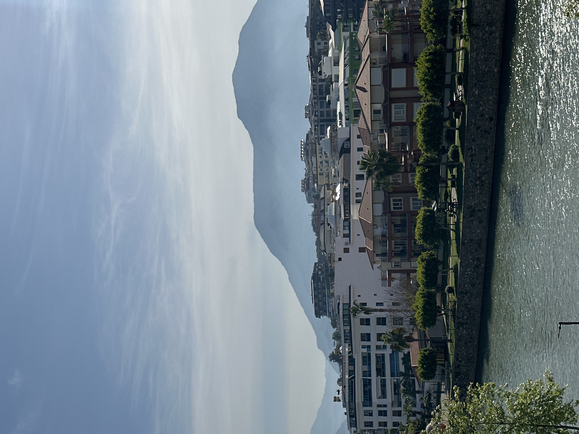 The image displays a panoramic view of a vibrant coastal or riverside town, consistent with the provided context of Alanya, Turkey. The scene, oriented horizontally, features distant mountains, a line of urban buildings, a waterfront promenade, and a body of water under a bright sky.
In the upper portion, the sky is a clear, light blue, adorned with delicate, wispy white clouds, indicating fair weather. Below the sky, a vast range of mountains stretches across the background. These mountains appear hazy and muted blue, suggesting they are quite distant and potentially obscured by atmospheric conditions.
The middle ground is dominated by a dense row of multi-story residential and possibly commercial buildings. Their architecture varies, with some structures featuring traditional reddish-brown roofs and balconies, while others exhibit more modern, stark white facades. Palm trees and other green foliage are interspersed among the buildings, adding greenery to the urban environment.
A distinct stone or concrete wall separates the buildings and their accompanying promenade from a large body of water on the right side of the frame. This water body, likely a river or the Mediterranean Sea, has a greenish-blue hue and is covered in ripples that reflect the bright sunlight, creating sparkling highlights. A prominent leafy green tree is visible in the immediate foreground, partially obscuring the water at the bottom right.
The time of day appears to be daytime, given the clear sky and strong sunlight. No human activity or discernible text is visible in the scene, which conveys a sense of peaceful urban and natural scenery.