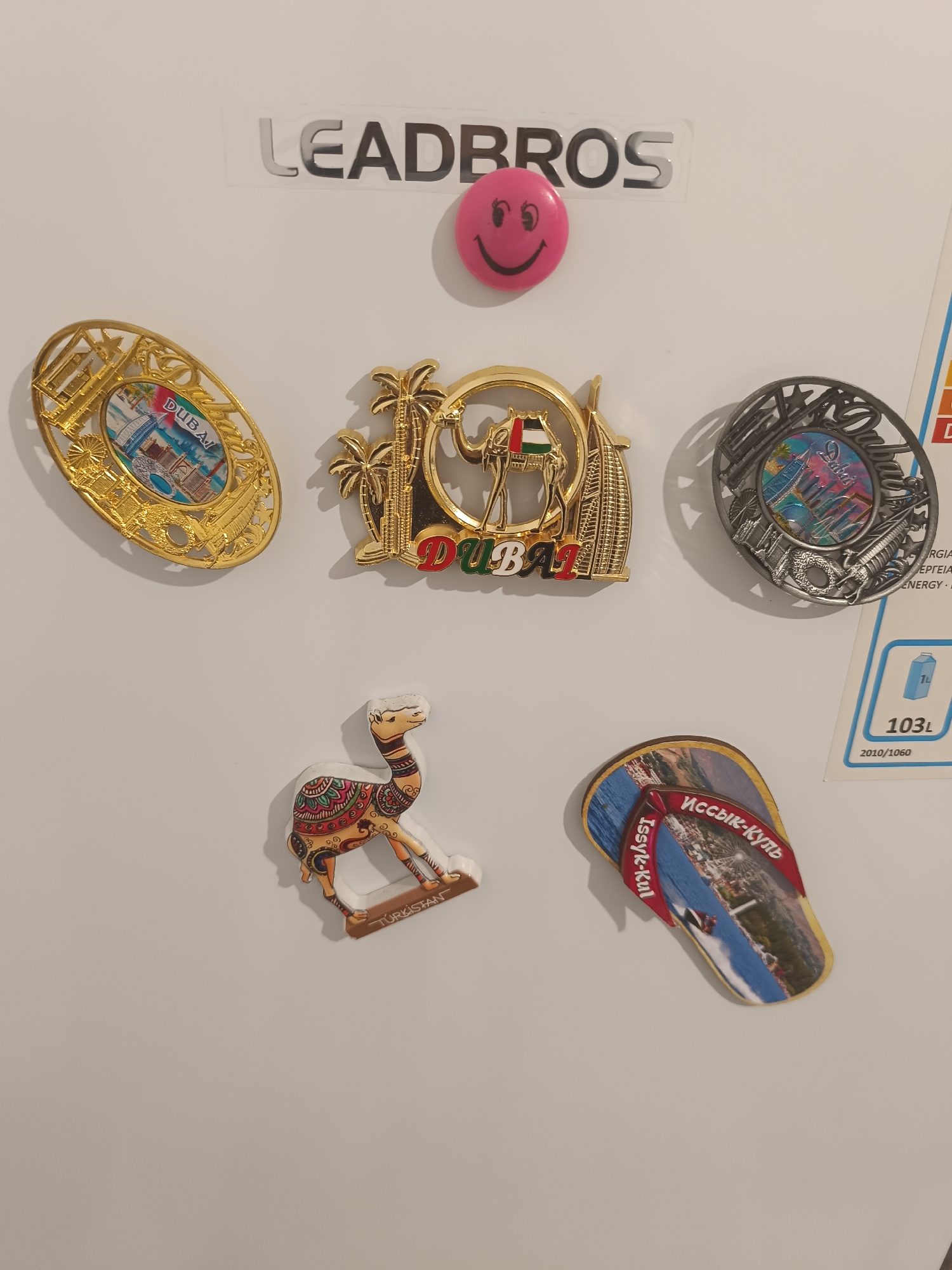 A close-up, eye-level, medium shot captures a white, vertical magnetic whiteboard adorned with various souvenirs. Dominating the top center is a rectangular sticker with black lettering spelling out "LEAD BROS". To the right of this text, a small, bright pink smiley face magnet sits.
Arranged in the upper half of the board are three circular magnets depicting Dubai landmarks. The leftmost magnet is gold-colored and features an intricate filigree design surrounding an image of buildings and the word "DUBAI". The central magnet is also gold and displays a camel with a flag, palm trees, and the word "DUBAI" in red. The rightmost magnet has a silver finish with a similar filigree pattern and an image of buildings.
In the lower half of the board, a camel-shaped magnet is positioned to the left. It is vibrantly colored with intricate patterns and has the word "TURKISTAN" written on its base. To the right of the camel, a flip-flop shaped magnet is displayed. The flip-flop features an image of a Ferris wheel and body of water, with text in a foreign script that appears to be "Issyk-Kul" and "Иссык-Куль" in red lettering.
A portion of a blue and white energy label is visible on the right side of the whiteboard, partially obscured by the souvenirs. The label includes text such as "ERGIA", "ΕΡΓΕΙΑΣ", "ENERGY", and a depiction of a milk carton with "1L" and "103L" printed on it. Below this label, the date "2010/1060" is visible. The overall environment is indoor, with consistent lighting illuminating the whiteboard and its contents.