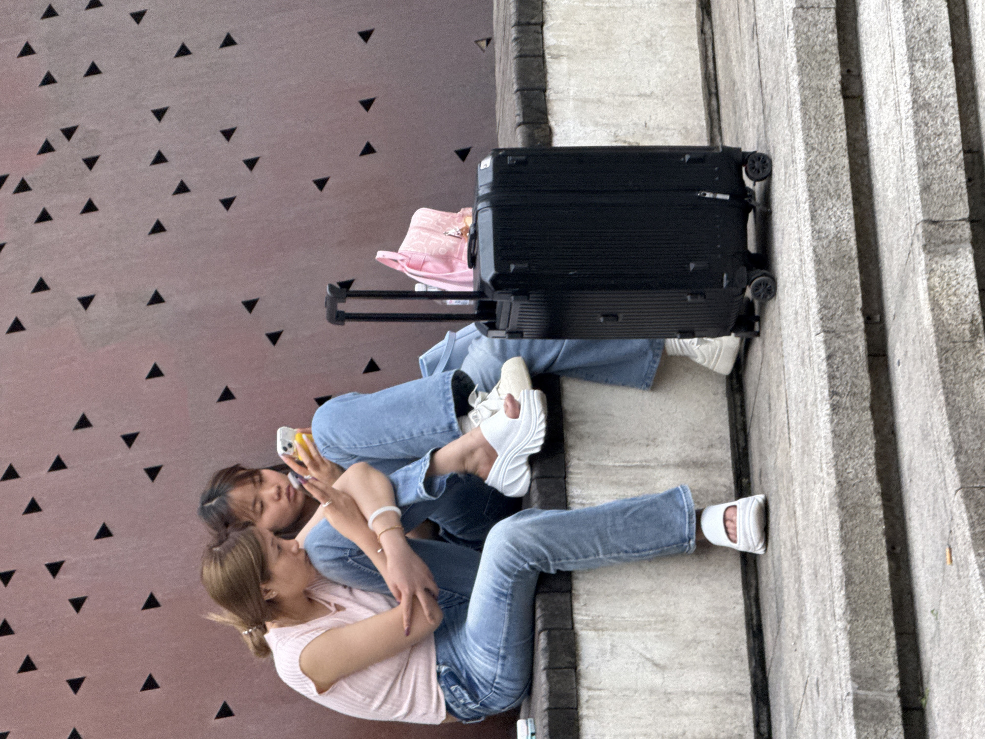 Two young women are sitting on a concrete surface next to a set of stairs in the Yuzhong District, China. A black suitcase and a pink sun hat are next to them. One woman is holding a phone.