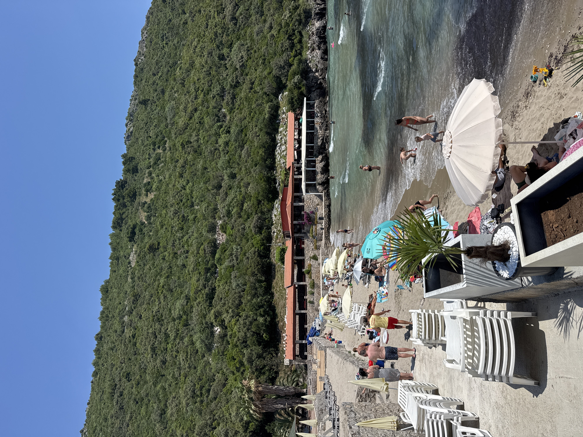 A sunny day at a coastal resort. A densely wooded, rocky hillside rises steeply behind a beach. The beach has a shoreline with light-colored sand and a calm, clear turquoise ocean. Several people are visible on the beach and in the shallow water.
On the beach, rows of stacked white lounge chairs are arranged on a concrete promenade. Some are set up with yellow umbrellas. A few people are walking on the sand, and others are sunbathing or sitting under umbrellas. A lifeguard wearing a yellow shirt and red shorts is walking across the sand.
A restaurant or beach club with a red-tiled roof and stone walls is situated near the back of the beach. The ocean has gentle waves. In the water, a few people are swimming or wading. A child is playing near the shoreline, and a young woman in a coral swimsuit is standing in the water. There are beach toys, including a yellow toy excavator, near the water's edge.
The overall scene depicts a relaxed and popular vacation destination on a clear day.