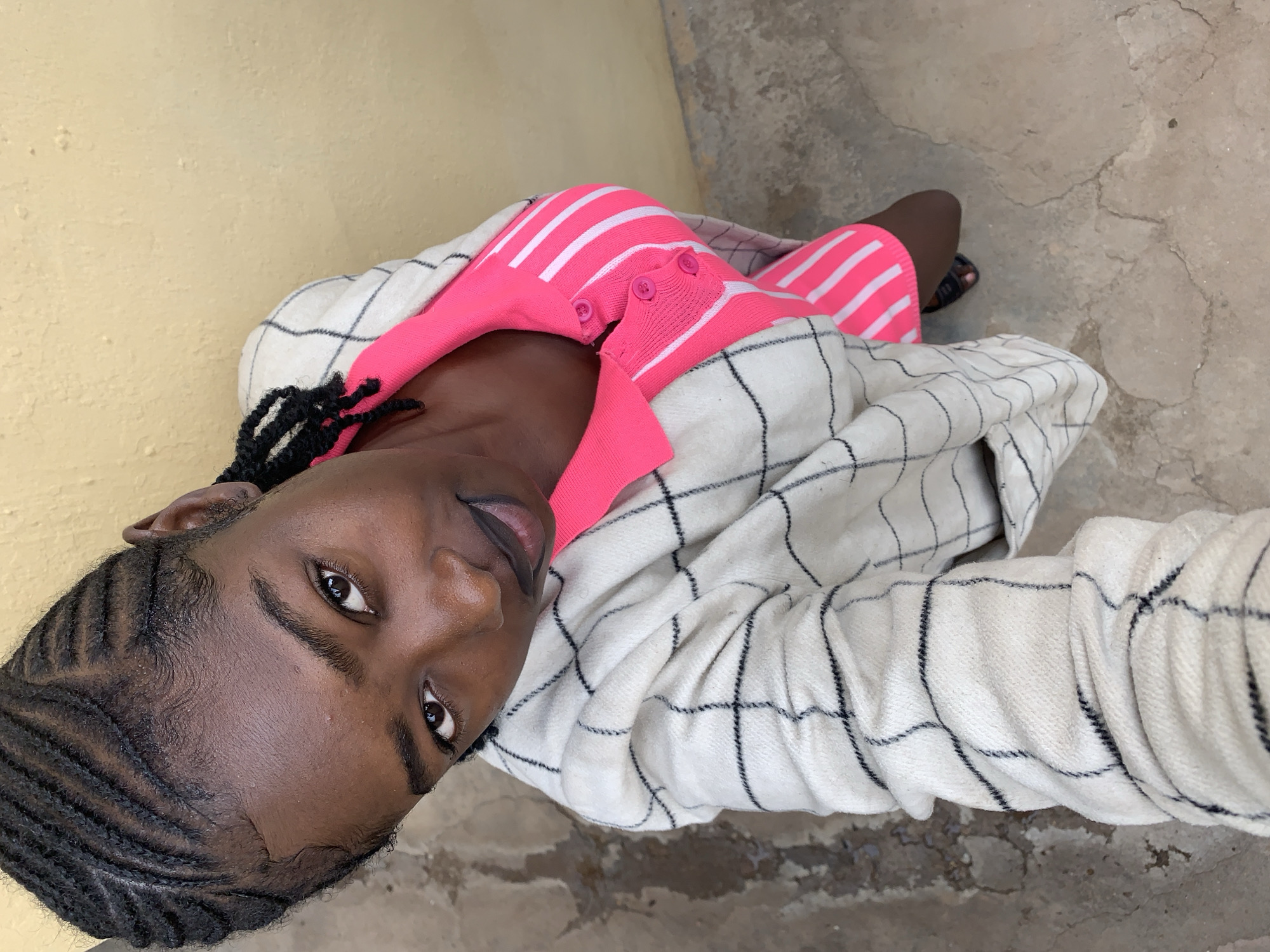A high-angle, top-down photograph in Rafin Par, Nigeria, captures a young Black woman looking directly upwards at the camera. Her head is positioned centrally, slightly tilted, with a direct gaze and a subtle smirk. Her hair is styled in tight black cornrows running in parallel lines across her scalp, with baby hairs defining her hairline. She wears a bright neon pink and white horizontally striped polo shirt with a collar and a three-button placket, visible under an off-white/cream, long-sleeved jacket featuring a black grid pattern. The fabric of the jacket appears textured or woven, with the sleeves slightly bunched. The background consists of a textured, light yellow/beige wall to her right and left, and an uneven, cracked concrete ground below, on which her left foot is partially visible, wearing a dark sandal. The lighting is bright and even, casting minimal shadows.