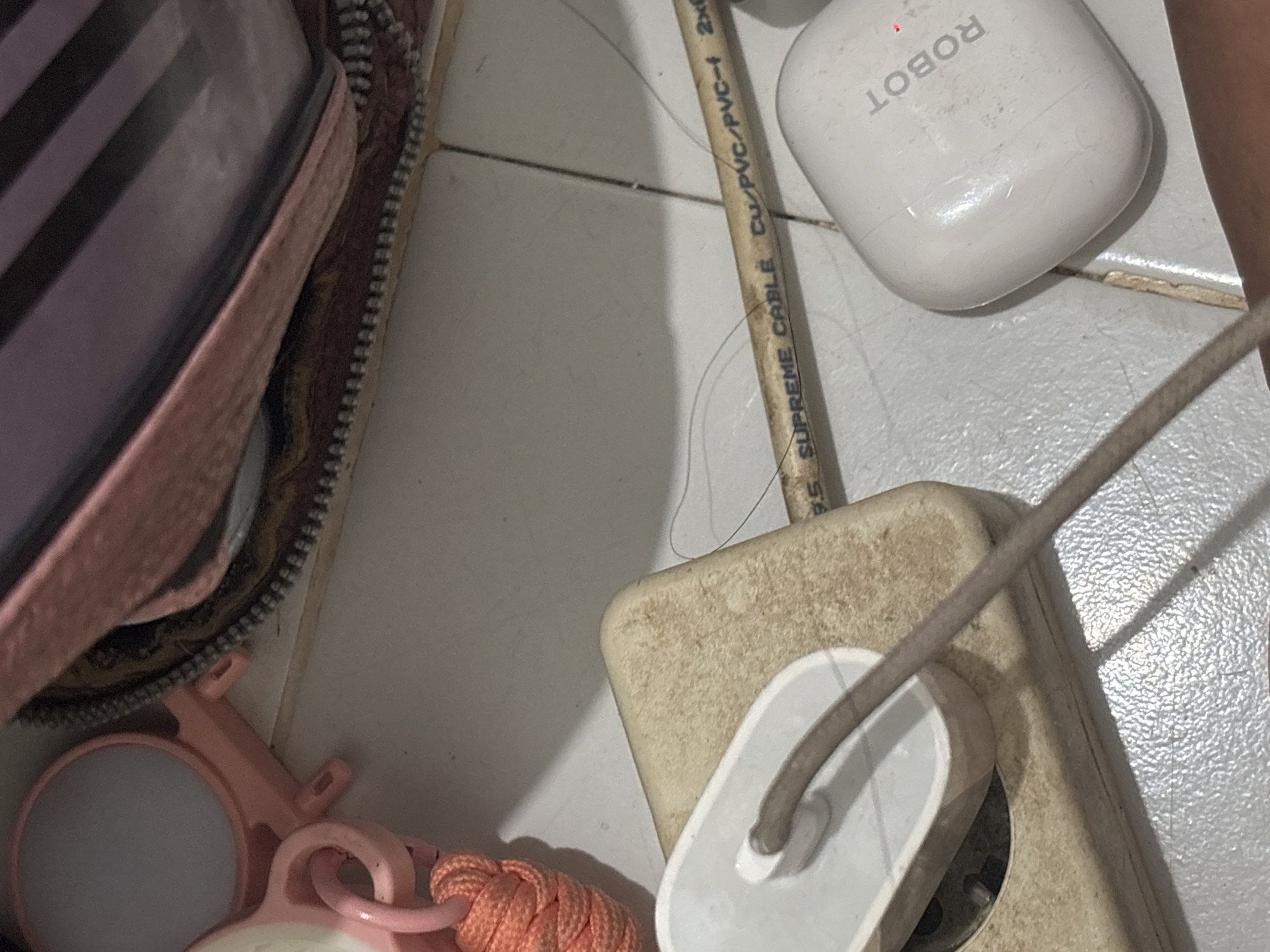 The image shows a close-up of electronic accessories on a tiled surface. In the foreground, a white electrical outlet is partially visible, with a white adapter plugged into it. A brown cord extends from the adapter and curves towards the right of the frame. To the right of the adapter, a white charging case for earbuds is present, with the word "ROBOT" printed vertically on its lid. A small red LED is illuminated on the charging case. A tan-colored cable, marked with "SUPREME CABLE Cu/PVC/PVC-f 2x", runs horizontally across the mid-ground of the image. In the background, a portion of a pink zippered pouch or bag is visible, along with a pink and orange lanyard or strap. The overall scene suggests a personal space, possibly a desk or floor in a home or office. The location in the city of North Jakarta, Indonesia, is not discernible from the image itself, as there are no specific geographical markers or contextual clues present.