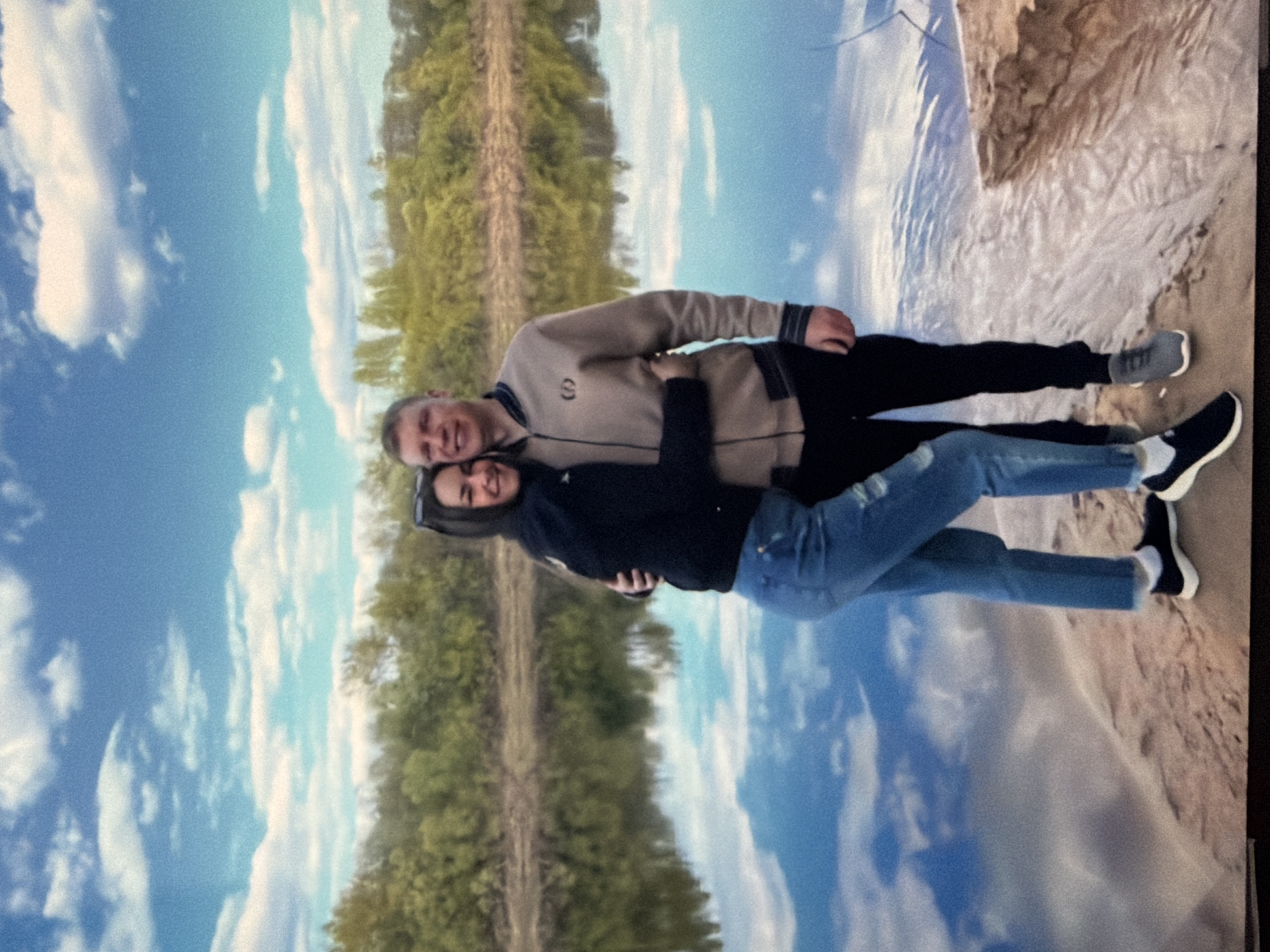 A man and a woman are posing for a photo in an outdoor setting. The man is wearing a tan jacket with a dark collar and cuffs and black pants. The woman is wearing a black jacket and blue jeans. They are embracing each other. In the background, there is a blue sky with clouds, a line of trees, and a sandy area. The location is Unknown, Unknown.