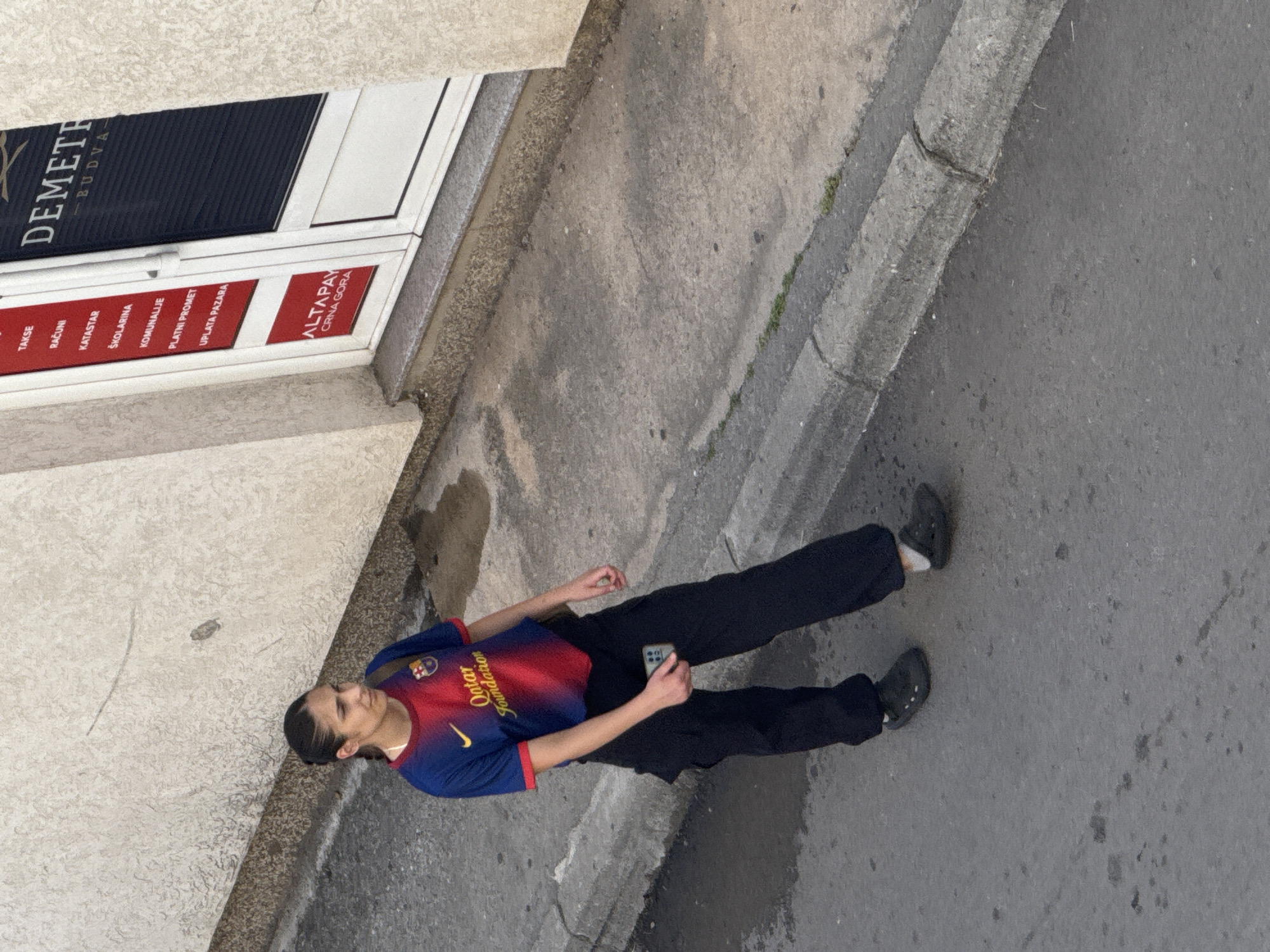 A person wearing a red and blue Barcelona FC soccer jersey and black pants stands on a concrete sidewalk next to a building. The person holds a smartphone in their hands. The building has a sign that reads "DEMETH - BUDVA" and "ALTAPAY CRNA GORA." There are several words listed in Serbian under the black sign, including "TAKSE," "RAČUNI," "KATASTAR," and "ŠKOLARINA." The location is the city of Budva, Montenegro. The person is wearing black Crocs.