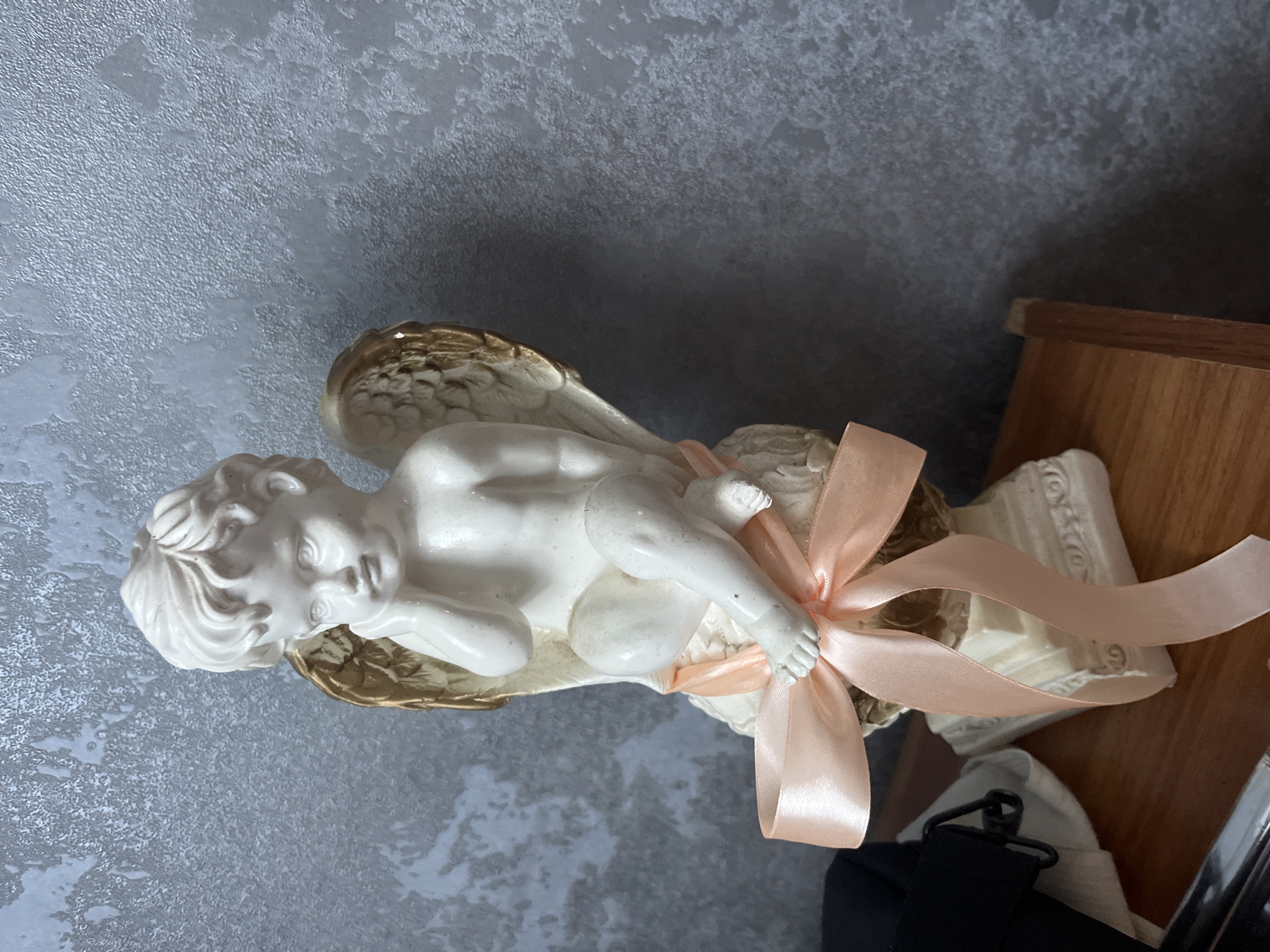 A white ceramic angel figurine with gold wings is seated on a pedestal. The angel has its right hand resting on its cheek, in a contemplative pose. A light pink satin ribbon is tied around its waist and over its legs, with a bow at the front. The figurine is positioned on a wooden surface, with a dark grey textured wall as the background. To the left of the figurine, a black bag and white fabric are visible. The scene is indoors, likely in a residential setting in Bohunove, Ukraine.