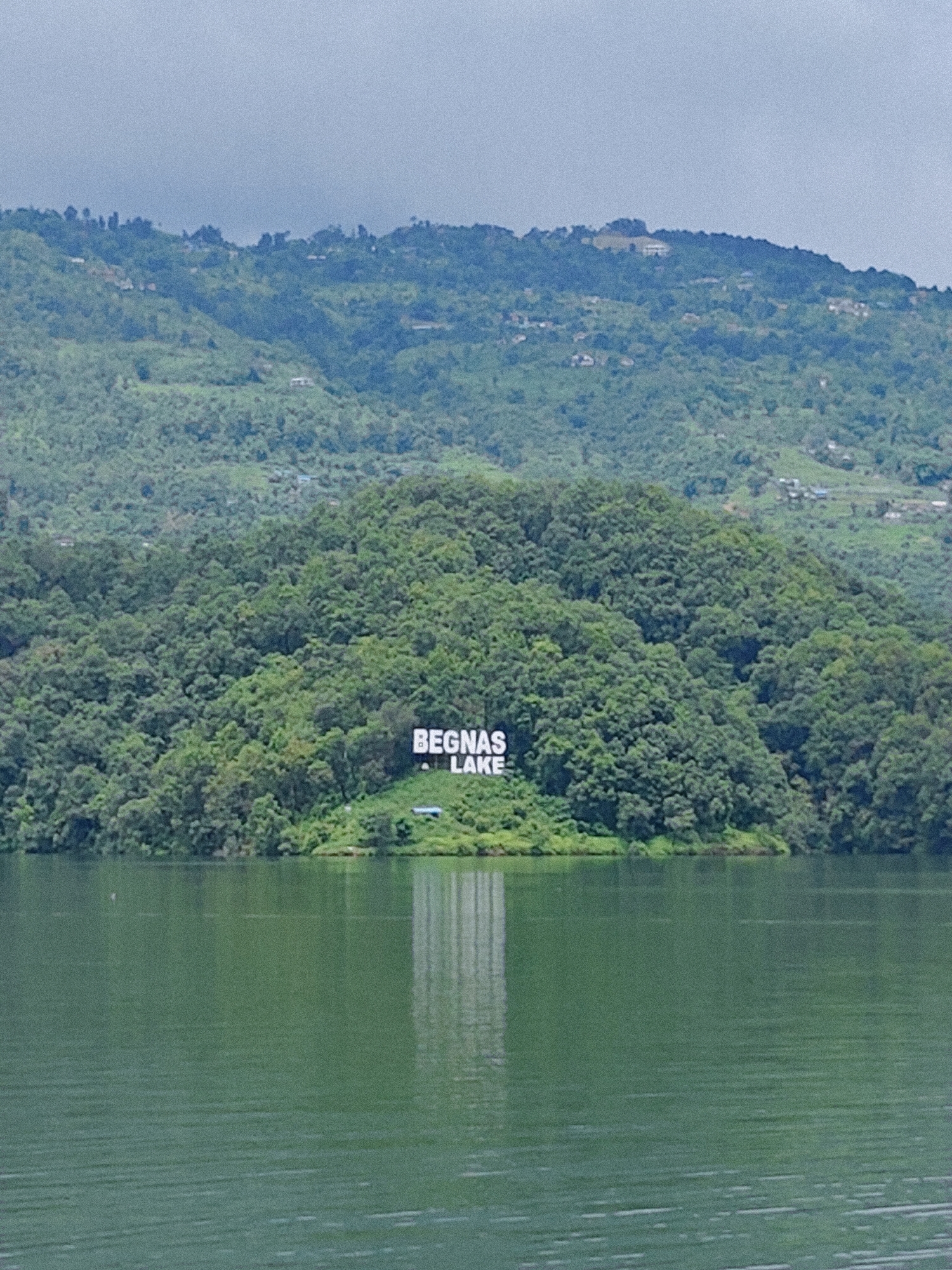 The image shows a wide shot of Begnas Lake in Pokhara, Nepal. In the middle ground, large white letters spelling "BEGNAS LAKE" are visible on a grassy incline of a forested hill. The lake itself occupies the foreground, appearing a deep green with subtle ripples on its surface. The background features rolling hills covered in dense green foliage, with some scattered buildings and a hazy sky above. The reflection of the letters and the hill is faintly visible in the water.