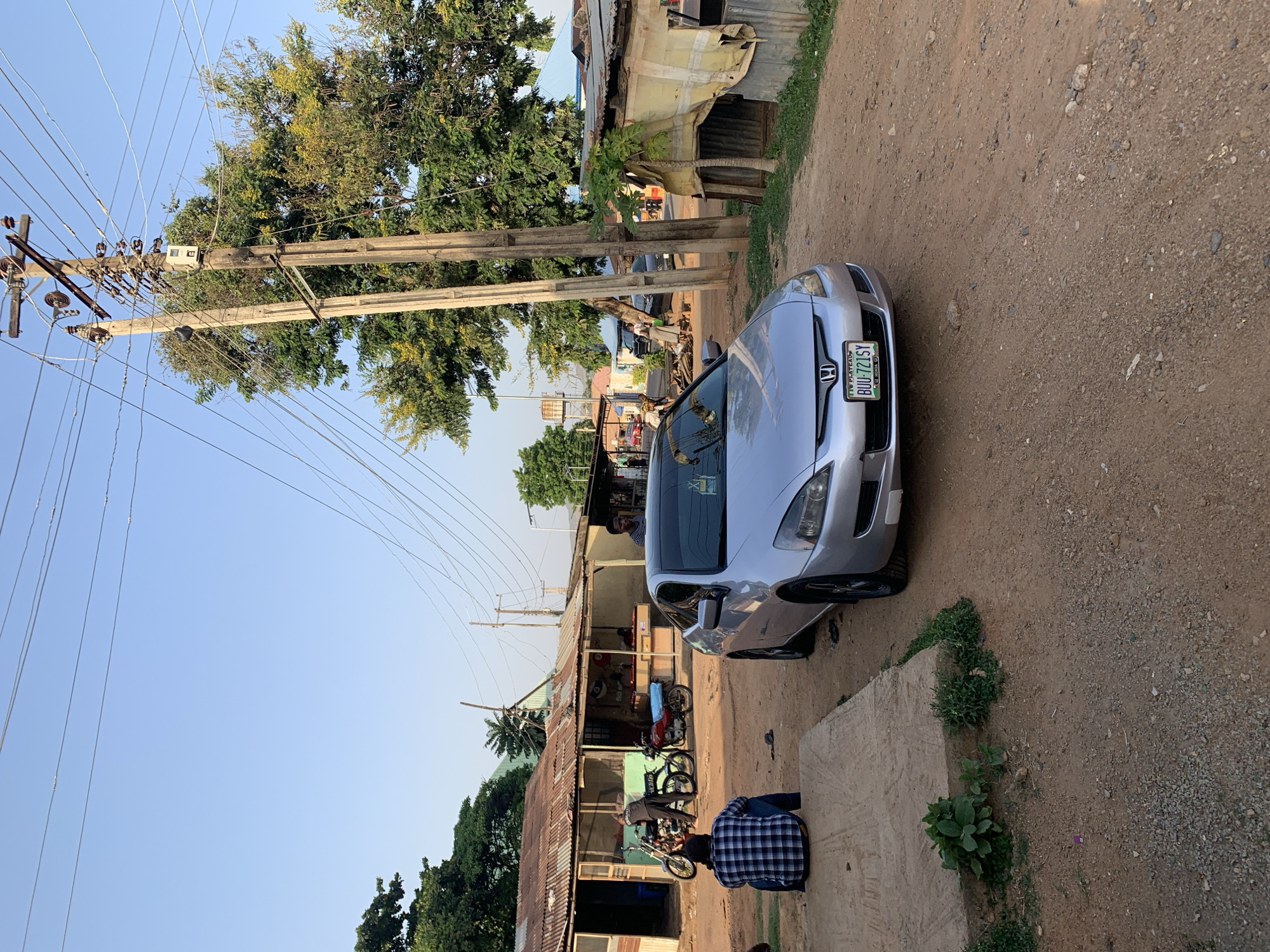A silver Honda sedan with a Nigerian license plate reading "BUU-721SY" is parked on a dirt road in what appears to be Kabong, Nigeria. The road is unpaved and dusty with visible rocks and pebbles. The car is situated next to a raised concrete surface. A person wearing a blue and white plaid shirt is kneeling on this surface. In the background, there are shops and structures, some with corrugated metal roofs. Several motorcycles are parked near the shops. Overhead, there are electrical poles and a tangle of wires. The sky is clear and blue, suggesting a bright, sunny day. Trees are visible throughout the scene. People can be seen standing and walking around in the area near the shops.