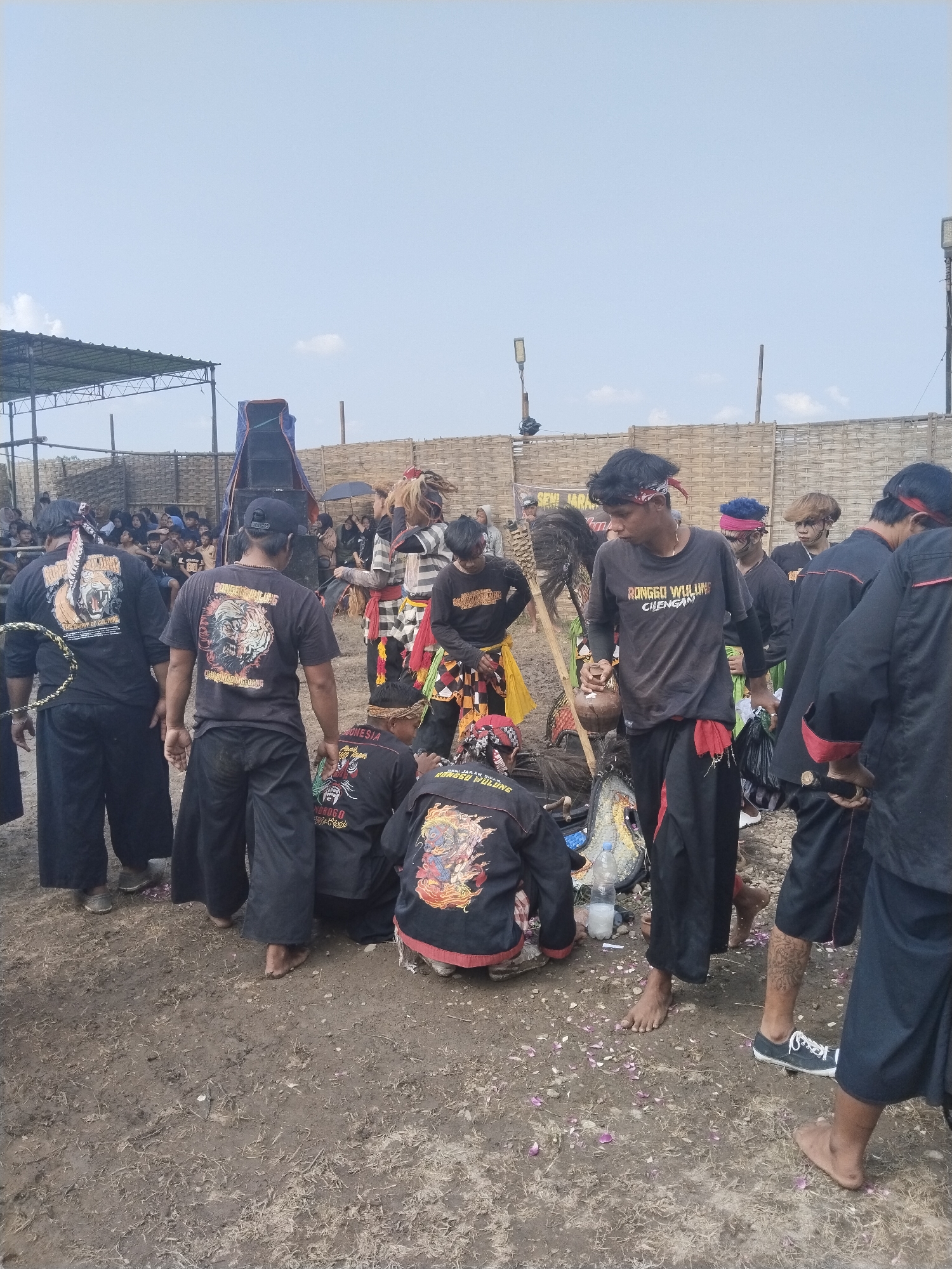 This outdoor image from Kedawung, Indonesia, captures a group of young men participating in what appears to be a cultural or traditional performance event during the daytime, under a clear sky with scattered clouds. The setting is a dirt field, with simple structures including a large metal-framed canopy on the left, a tall speaker covered with a blue tarp, and a woven bamboo fence forming the background.
The main subjects are numerous young men, predominantly dressed in black t-shirts and dark, loose-fitting pants, often accessorized with colorful sashes or headbands. Many of their t-shirts feature vibrant graphic designs, often depicting mythical creatures or fierce animals like tigers. Visible text on their clothing includes "THE SPIRIT OF CULTURE," "RONGO WULUNG CIREBON," and "INDONESIA." A partially visible banner in the background reads "SENI JARANAN," indicating a Jaranan (horse dance) performance, a traditional Indonesian art form.
The men are engaged in various activities: some are standing and conversing, others are kneeling or bending down, possibly arranging costumes or props. One man on the right holds a traditional coconut shell vessel, while a plastic bottle lies on the ground near others. Scattered pink and white rose petals are visible on the dirt, often used in ceremonial contexts. Some participants are barefoot, while others wear shoes. In the background, beyond the main group, spectators are gathered, some taking shelter under umbrellas. The scene conveys an atmosphere of preparation or a break during a communal cultural celebration.