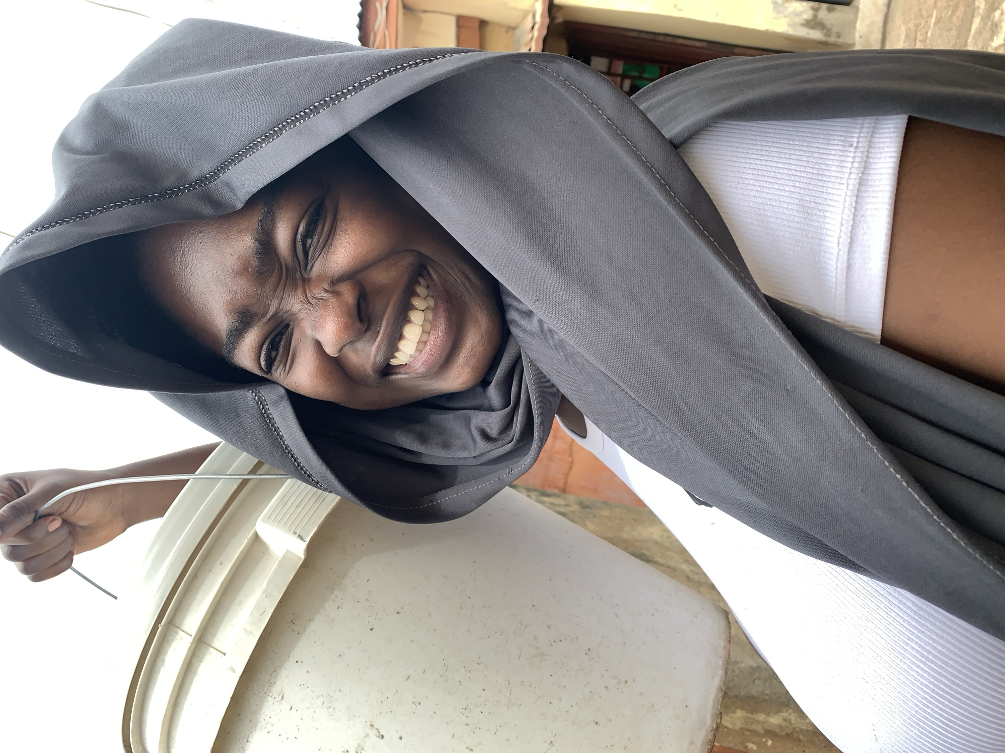 A young woman smiles at the camera, wearing a gray hooded garment over a white ribbed tank top. Her head is tilted. She stands near a large white plastic bucket. A metal rod extends from her hand towards the bucket. The background suggests an outdoor, perhaps rural, location, possibly within or near Rafin Par, Nigeria. Building elements are visible in the background.