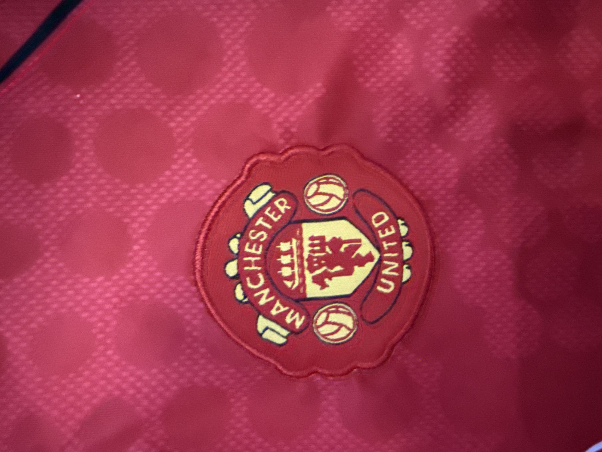The image shows a close-up of the Manchester United Football Club crest, embroidered onto a red fabric. The crest features a shield with a red devil in the center, a ship above it, and a football on the left. The words "MANCHESTER" and "UNITED" are written in yellow lettering around the crest. The fabric has a subtle, repeating pattern of small diamond shapes. There is no indication of the city of Burgas, Bulgaria, in this image.