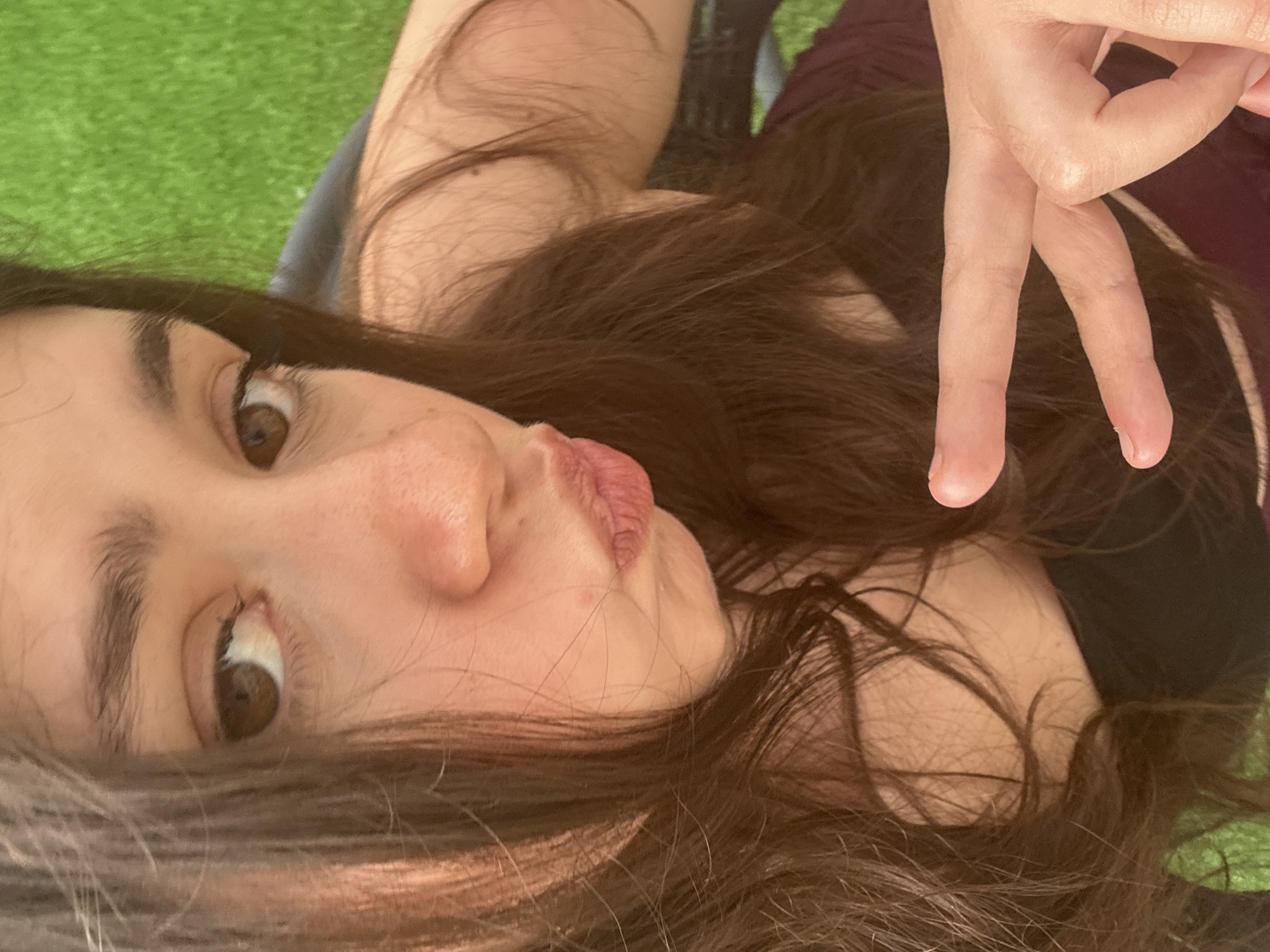 This image presents a close-up, high-angle portrait of a young woman with fair skin, long dark brown hair, and dark brown eyes. She is looking directly towards the camera, her lips pursed in a pout. Her hair, styled with a slight wave, cascades around her face and over her shoulders, partially obscuring the right side of her face. Her right hand (viewer's left) is prominently featured in the foreground, forming a peace sign with two fingers extended. She is wearing a dark-colored, possibly strapless, top, with a segment of a reddish-brown or burgundy garment visible beneath her arm. The background consists of a brightly lit, out-of-focus green area, indicative of grass, suggesting an outdoor setting. A dark, indeterminate object, possibly a piece of furniture, is partially visible behind her right arm. The lighting appears to be natural and bright, illuminating her face and the highlights in her hair. The specific geographical location, such as Garmen, Bulgaria, is not discernible from the visual information present in the image.