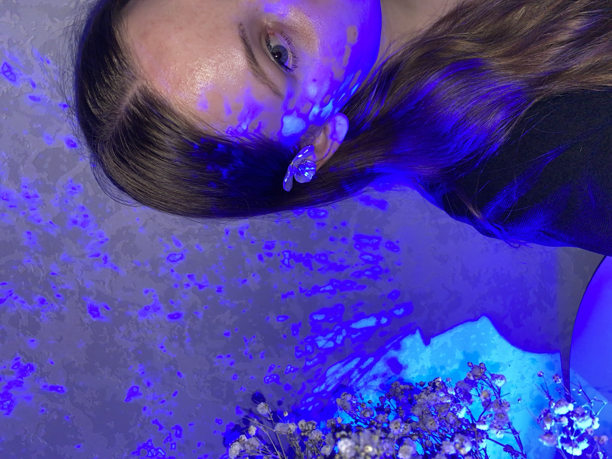 A person is depicted in an indoor setting under blue lighting. The person's face, turned towards the viewer, shows a single eye and part of their forehead. They wear a floral earring. Their hair is brown and styled back. In the lower portion of the image, a bouquet of baby's breath flowers is visible. The background appears to be a mottled surface, likely a wall, also illuminated in blue. There is no indication of the city of Izhevsk, Russia, in the image.