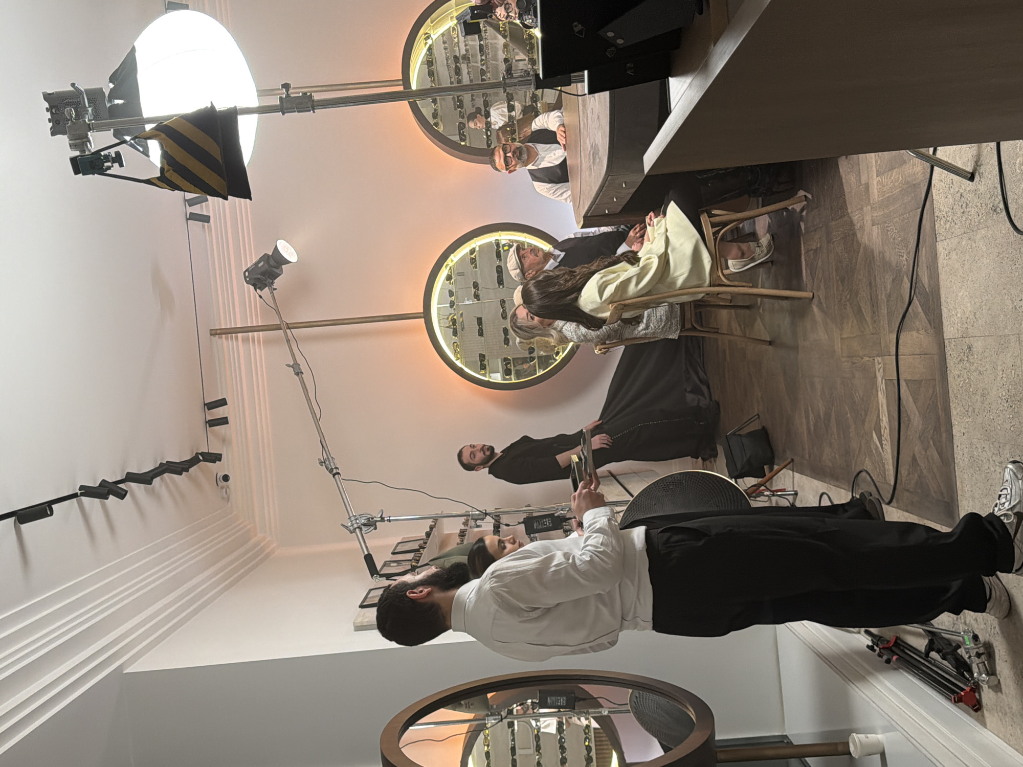 The image depicts a film or photography set indoors, likely in a retail space, possibly an eyewear store. A man in a white shirt and black pants appears to be working on set, holding a reflector or monitor. Several people, including at least two seated customers, are situated at a counter; a man with glasses is standing behind the counter. The store has round mirrors displaying eyewear. There is film equipment visible: lights on stands and booms. The location is Yerevan, Armenia, though this is not readily discernible from the image alone.