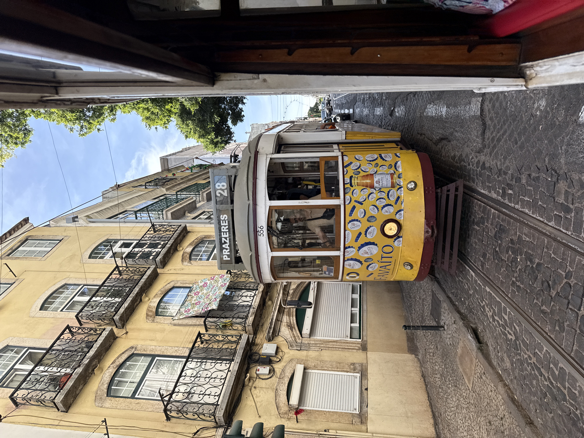 This image shows a yellow tram with the number 28 and "PRAZERES" written on it, on a cobblestone street in Lisbon, Portugal. The tram is decorated with images of bottle caps and a bottle of Favaito. The tram is passing by a yellow building with balconies and arched windows. The street is wet, suggesting it might have rained recently or has been washed. Trees and buildings are visible in the background, with a blue sky and some clouds. The image appears to be taken from another tram or a close vantage point.