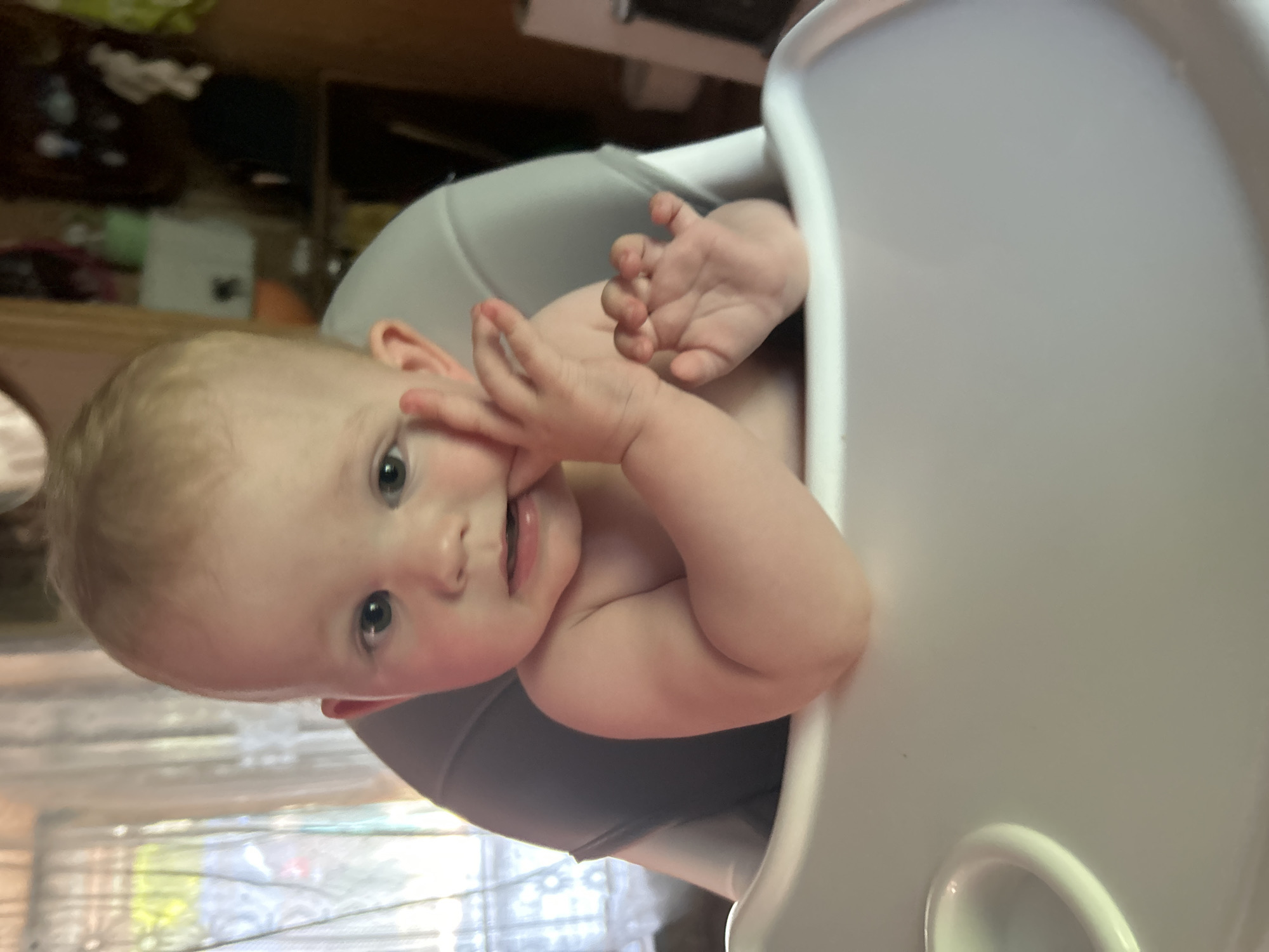 A baby with blonde hair and blue eyes is sitting in a high chair in what appears to be a home environment. The baby is shirtless and has their fingers in their mouth. The high chair is white with a grey insert. In the background, there is a window with sheer curtains and some shelves with items on them. The lighting suggests it is daytime. The exact location in Ozeretskoe, Russia, cannot be determined from the visual information provided.