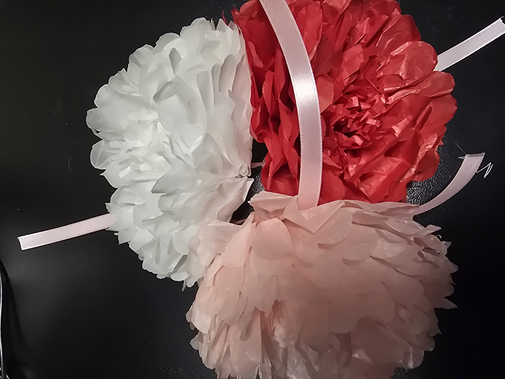 Three tissue paper pom-poms, one white, one pale pink, and one red, are arranged on a dark, reflective surface. Each pom-pom is tied with a thin, pale pink ribbon, with the ribbons extending outwards from the pom-poms and some crossing over each other. The pom-poms appear to be decorative or party items.