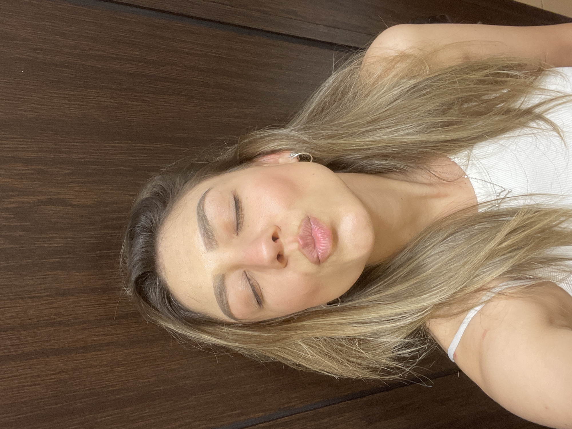 A light-skinned female individual, appearing to be in her 20s or 30s, is depicted in a close-up, chest-up shot, likely a selfie. Her eyes are closed, and her lips are pursed in a puckering or kissing gesture, with a slight tilt of her head to her right (viewer's left). Her long, straight to wavy hair, featuring light brown and blonde highlights, cascades over her shoulders and chest. She is wearing a white, ribbed tank top or camisole. A thin, silver-colored chain necklace with a small, geometric pendant is visible around her neck, and small, silver-colored hoop earrings adorn both earlobes. Subtle reddish marks are present on her left shoulder. The background is a dark brown, vertically textured surface, indicative of wood paneling, a door, or furniture. The scene is indoors, in the city of Unknown, Unknown.