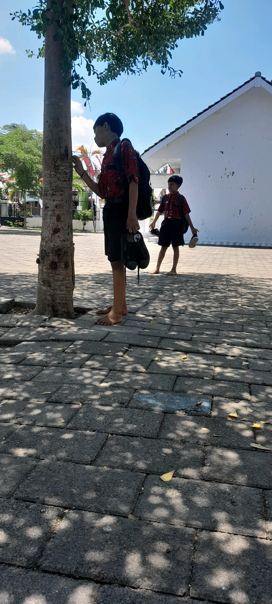 Two young boys, likely students due to their uniforms, are outdoors on a sunny day. The boy in the foreground, closest to the camera and a tree trunk, wears a red patterned shirt, black shorts, and a black backpack. He is barefoot and holding a pair of black shoes in his right hand. He appears to be interacting with the tree. Behind him, further into the scene and slightly to the right, is another boy in similar attire, also barefoot and carrying a pair of black shoes. He is walking away from the camera. They are on a paved area made of interlocking stone blocks, with shadows from overhead foliage dappling the ground. A white building with a tiled roof is visible in the background. The environment suggests a school or public space in a warm climate. The presence of Indonesian flags in the background suggests the location is Indonesia, possibly Rogojampi.