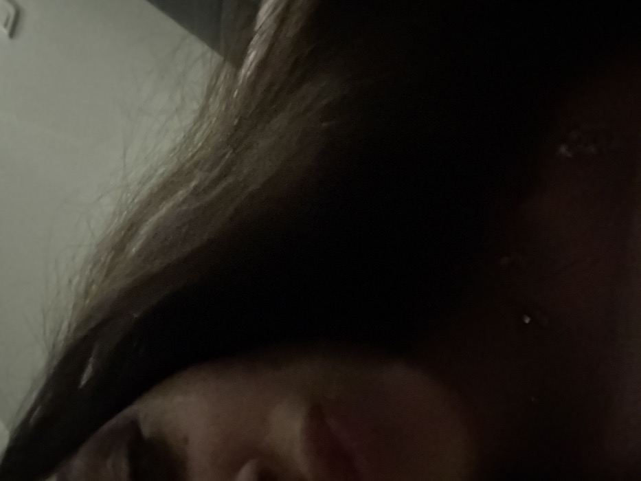 A close-up shot focuses on a face, with a partial view of the nose and the corner of an eye visible. Dark hair dominates the upper portion of the image, obscuring much of the background. The lighting is dim, casting shadows and limiting visibility. The overall composition is intimate, capturing a personal and partially obscured view of the subject.