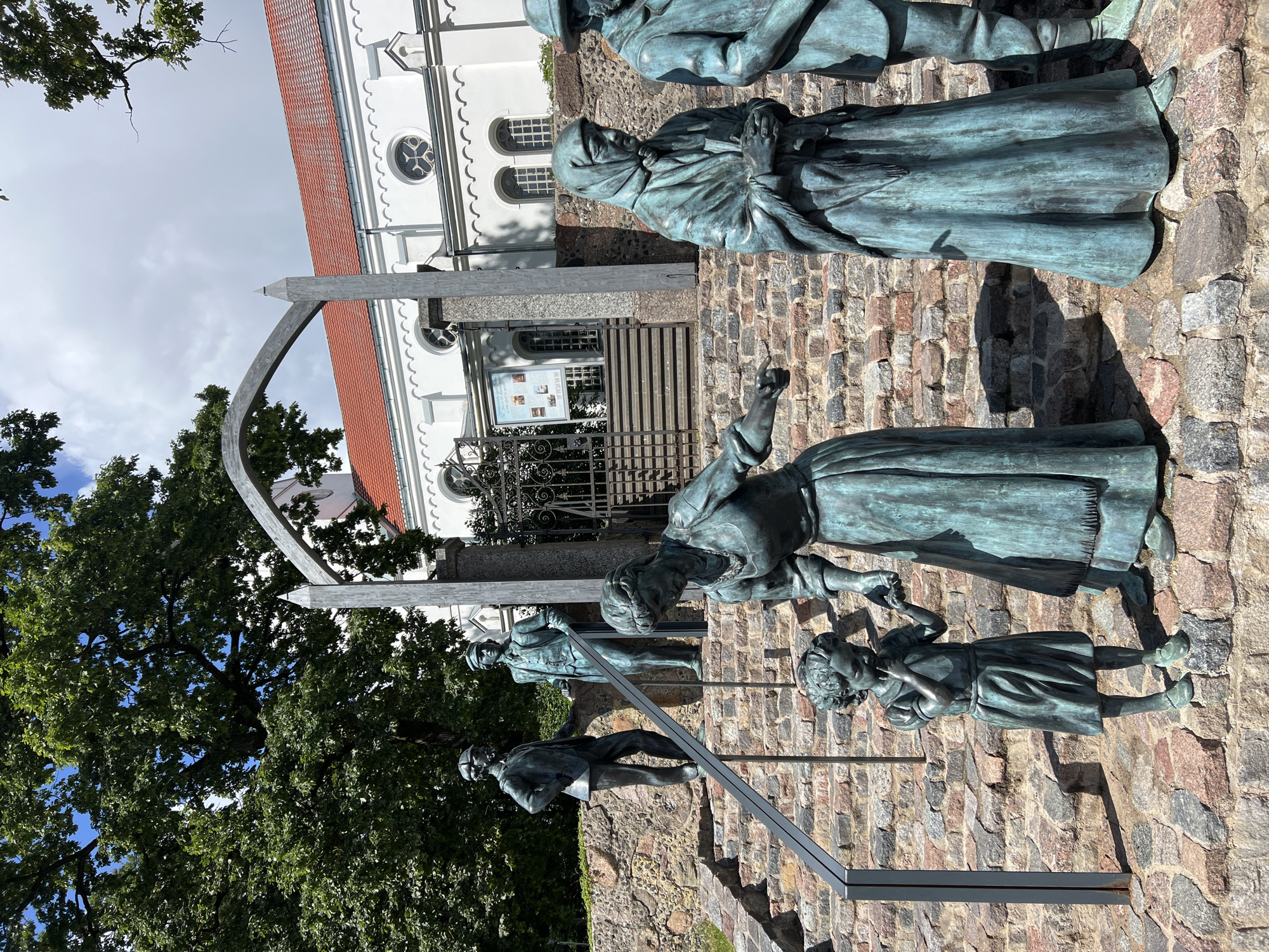 The image depicts a bronze sculpture depicting a group of people on a stone staircase in front of a white building with a red-tiled roof. The sculpture is located in the city of Saldus, Latvia, specifically at the Saldus Historical Museum. The main figures in the sculpture include an adult female with a child standing at her side, and two other adults in the background. The adult female in the foreground is leaning towards the child, who appears to be touching their mouth. Another adult figure, also in historical attire, stands to the right of the child, holding something in their hands and looking down. Further back, two male figures in suits and hats are positioned on the stairs. Behind the sculptures, a gateway with an arch leads to the entrance of the building, which features arched windows and decorative architectural elements. The environment is outdoors, with a large tree with lush green foliage visible on the left side of the image, and a cloudy sky above. The stone staircase and the surrounding walls are constructed from rough-hewn stones of various sizes and colors. The bronze figures show signs of verdigris, indicating their age and exposure to the elements.