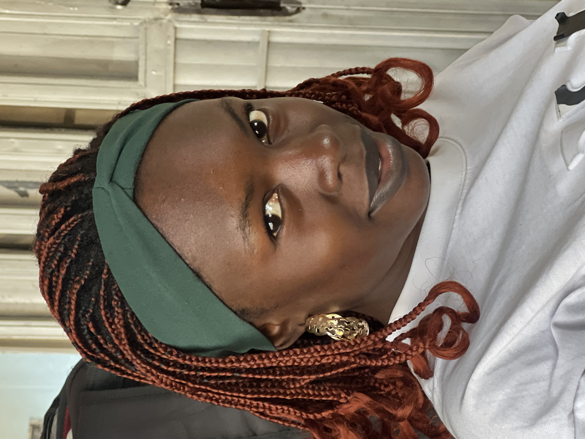 A close-up, head-and-shoulders portrait shows a young Black woman with braided, reddish-brown hair and a dark green headband. She is wearing a white sweatshirt and large gold hoop earrings. Her skin is dark and shiny, and she is wearing dark lipstick. Her gaze is directed towards the camera. The background consists of a plain, light-colored wall and a doorway with white doors. The lighting is natural and comes from the front. The image appears to have been taken outdoors in an urban setting, possibly in Jos, Nigeria, given the context provided.