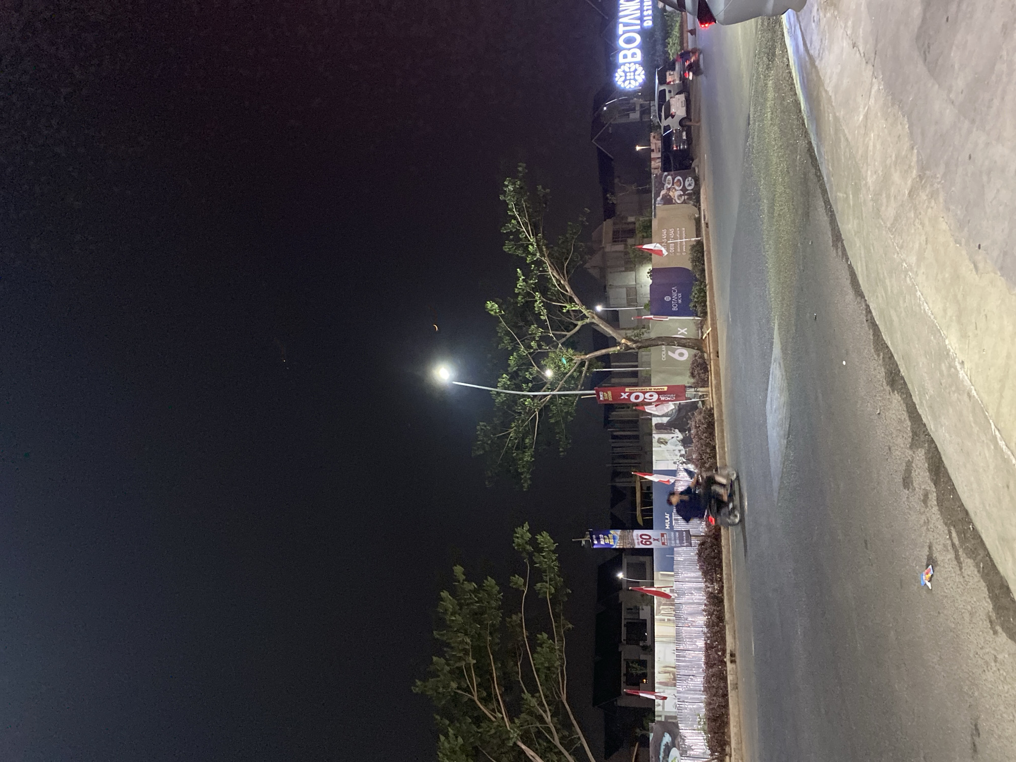 This is a nighttime, eye-level, medium shot of a street scene in South Tangerang, Indonesia. In the foreground, a paved road curves towards the right of the frame, with a concrete sidewalk on the right edge. A small, colorful object, possibly a discarded wrapper, lies on the asphalt. In the mid-ground, a motorcycle with two occupants is traveling away from the camera on the left lane of the road. On the left side of the road, there are trees with green foliage. A row of flags, predominantly red and white, are planted along a construction fence. Several banners are visible, including one that reads "60". Beyond the fence, houses are under construction. On the right side of the mid-ground, there are parked cars, including a white SUV and a black sedan, with a motorcycle in front of them. A large illuminated sign on the far right reads "BOTANICA DISTRICT". The sky is dark, with a faint crescent moon visible near the top left. Streetlights illuminate parts of the road and the area around the "BOTANICA DISTRICT" sign.