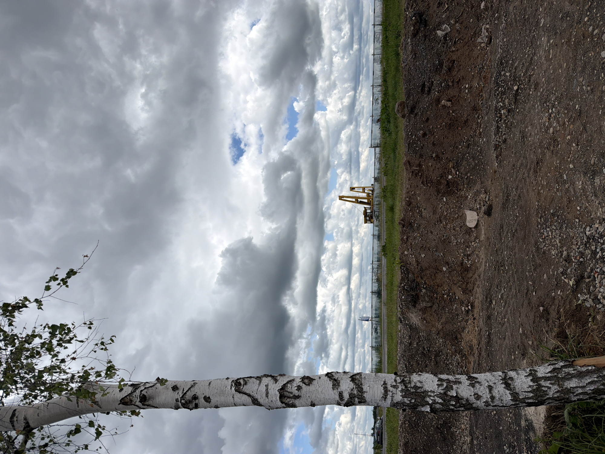 The image depicts an outdoor scene with a birch tree in the foreground, partially obscuring a construction site. The construction site features yellow machinery and a fence line. The sky is filled with dramatic, gray clouds, with a few patches of blue sky visible. The ground in the foreground is a mix of soil and gravel, suggesting an unfinished or disturbed area. In the middle ground, there is a strip of green grass and the aforementioned fence. A road with a vehicle is visible in the distance, along with a radio tower. The overall impression is of a partly overcast day at a developing or under-construction location. The location is identified as Папярнянскі сельскі Савет, Belarus.