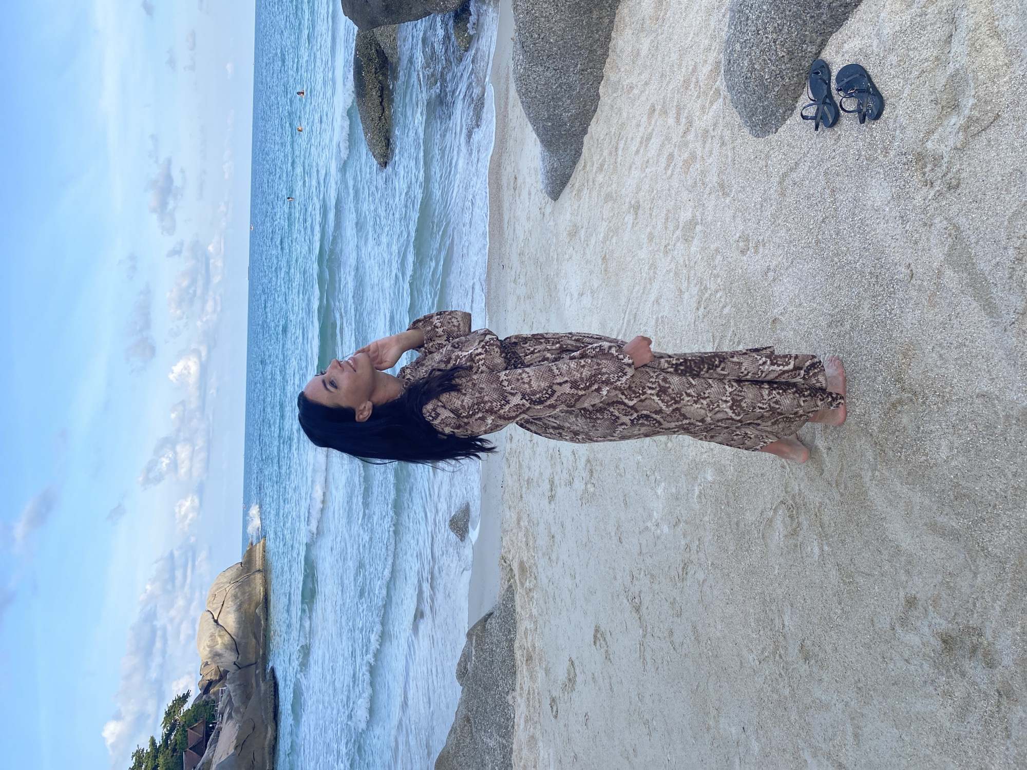 The image depicts a woman in a long, brown patterned dress standing on a sandy beach next to the ocean in Ban Haad Rin, Thailand. She is looking upwards, with her hand near her face. To the left of the frame is a rock formation next to a building with trees, and to the right of the frame are other rocks with a pair of black flip-flops on the sand. The sky above the ocean is partly cloudy.