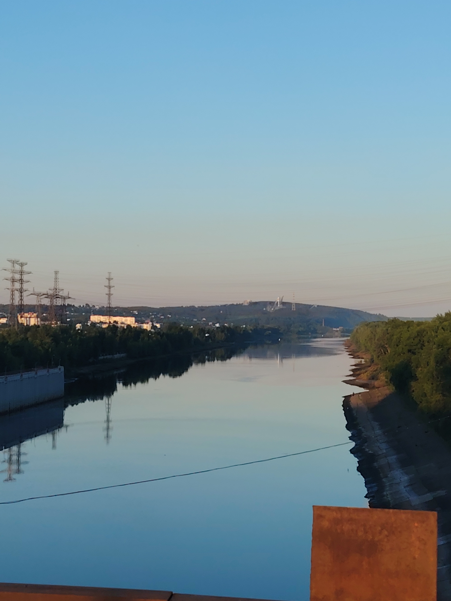The image shows a wide, calm river with its surface reflecting the clear, pale blue sky and the surrounding landscape. On the left bank, a concrete embankment is visible, followed by a dense line of green trees. Beyond the trees, a city skyline emerges, featuring several tall, lattice-like transmission towers and multi-story buildings. The right bank of the river is lined with trees and a sloped, rocky embankment. In the distance, a hazy, undulating hill is dotted with more transmission towers and some structures. The water is smooth, creating a mirror-like reflection of the sky and the scene on both banks. The overall impression is one of a serene, possibly early morning or late evening scene in an urban or semi-urban area. The presence of transmission towers and industrial-looking buildings suggests a location with a power infrastructure.