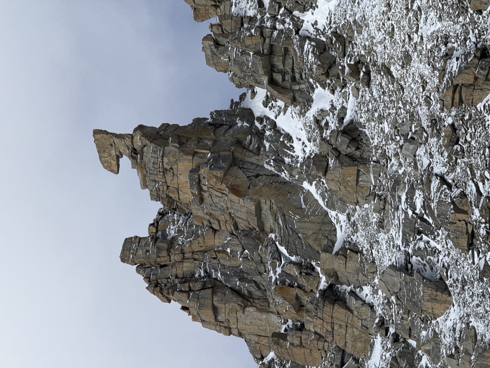 A vertically oriented image captures a dramatic, rugged mountain face composed of heavily fractured and layered rock formations. The rock, predominantly dark grey and brown, is extensively covered with white snow and ice, clinging to ledges, crevices, and vertical surfaces. Icicles are visible in several areas, indicating freezing temperatures.
The mountain face occupies the right and lower sections of the frame, contrasting with a clear, light blue to grey sky that dominates the upper and left portions. A prominent, irregularly shaped rock formation juts out sharply from the main cliff on the upper left, resembling a natural cantilever or a weathered peak.
No people, animals, or man-made structures are visible, underscoring the raw, untamed nature of the environment. The scene depicts a cold, high-altitude landscape, likely during winter or early spring due to the substantial snow and ice cover. The lighting suggests it is daytime. This mountainous terrain is located near Parga town, China. There is no visible text within the image. The overall impression is one of stark natural beauty and geological resilience.