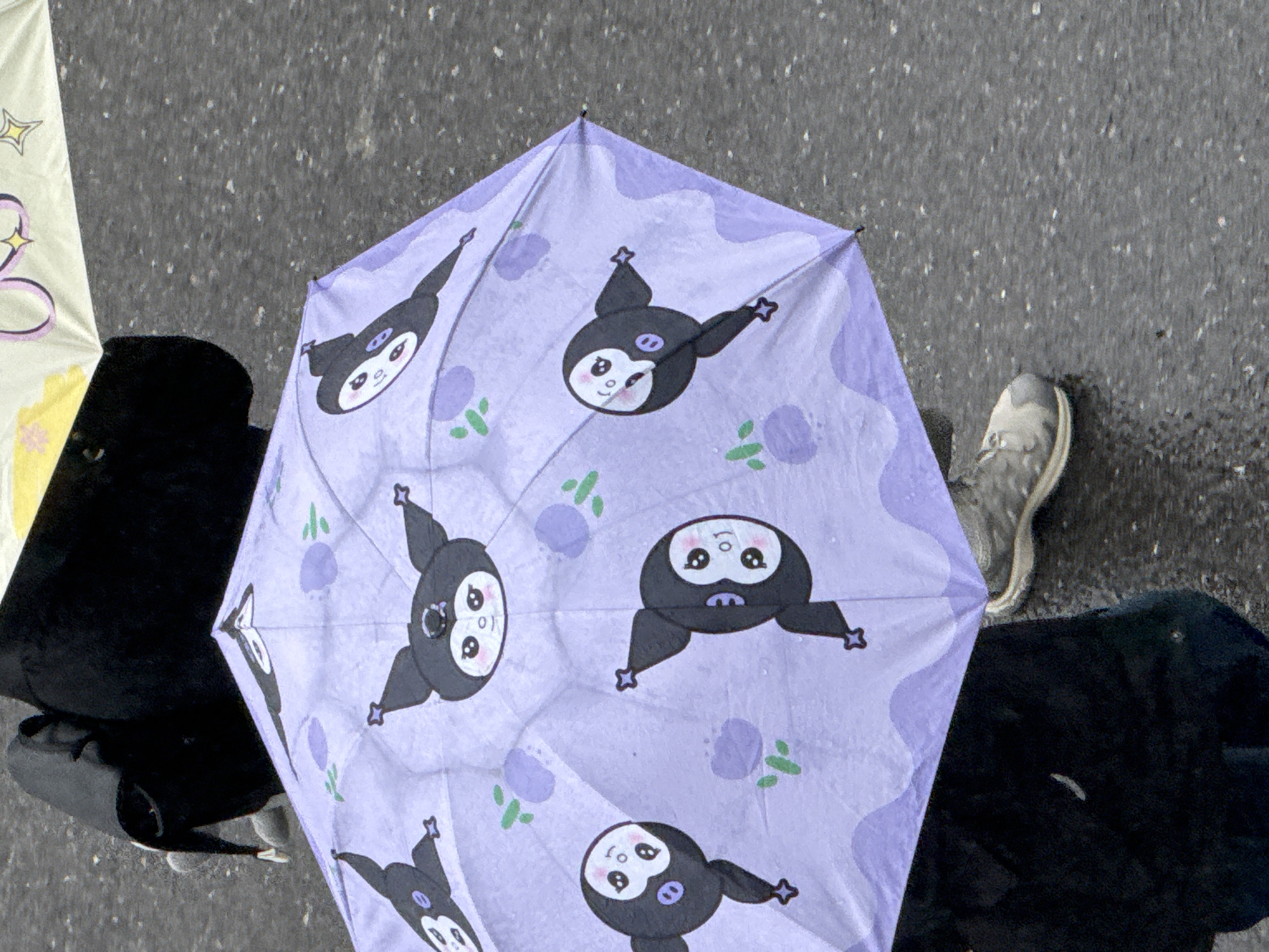 An overhead shot shows a lavender umbrella with a cartoon character pattern, next to a black backpack and a gray sneaker on wet pavement. The location is presumed to be Zhongtun, China.