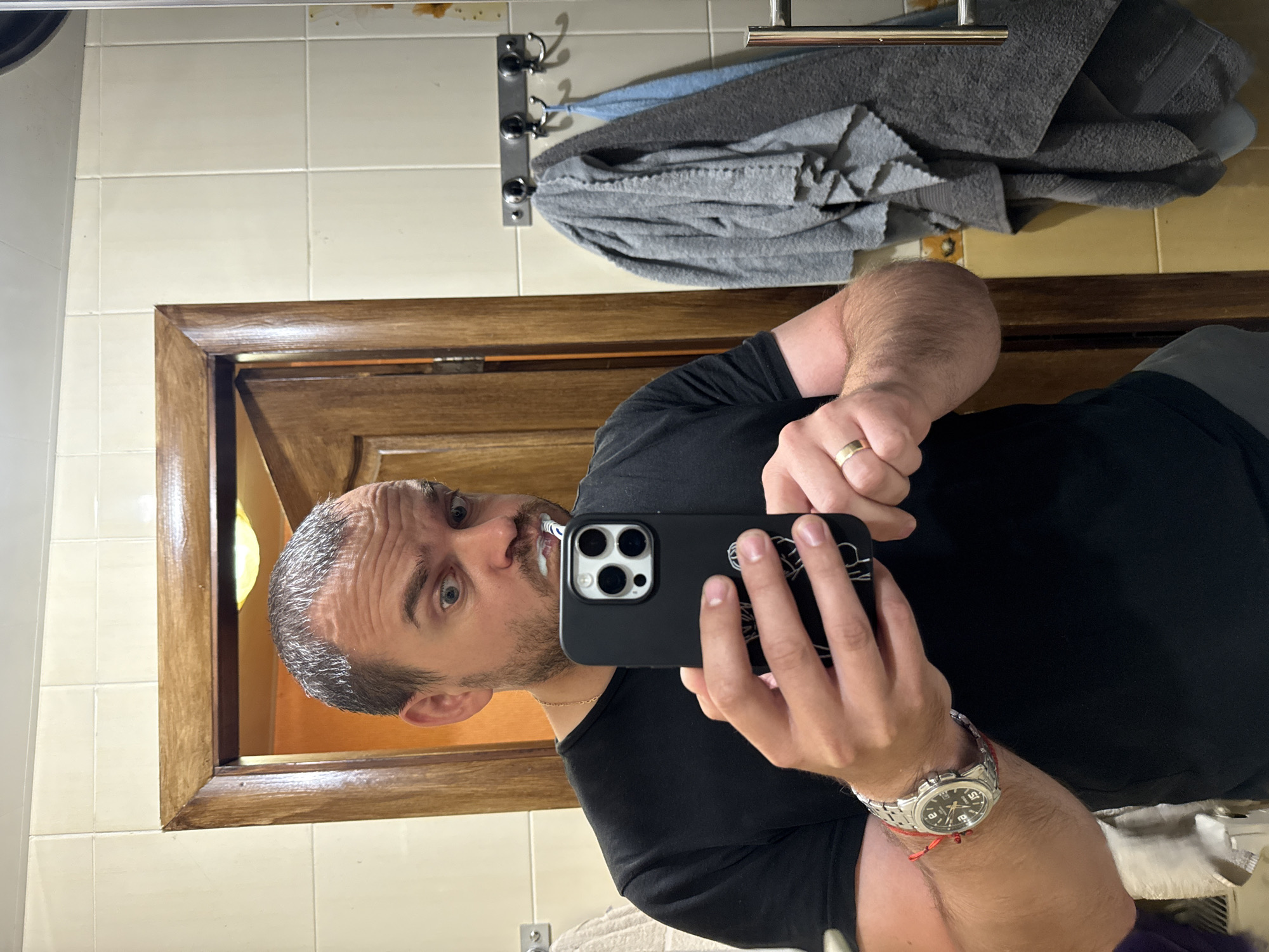 A man is taking a selfie in a bathroom, holding a phone in his right hand and a toothbrush with toothpaste in his mouth. He is wearing a black t-shirt, a silver watch on his left wrist, and a gold ring on his right ring finger. The bathroom appears to be in a private residence, likely a home bathroom, not a public facility. The background shows a wooden door, tiled walls, and several towels hanging on hooks. The overall impression is of a candid moment captured in a personal space. There is no information in the image to suggest it is located in the city of Valky, Ukraine.