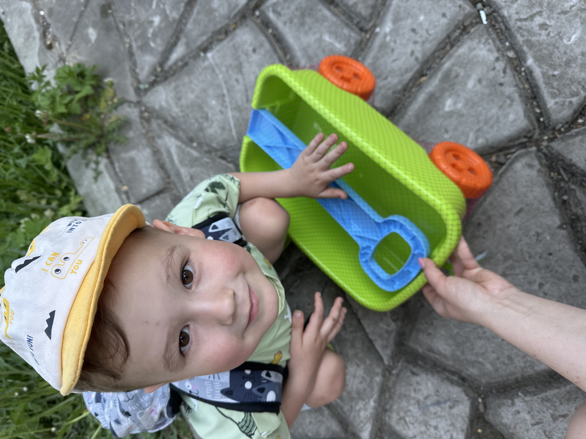 A young child with a tan cap and a light green shirt sits on a gray paved surface, with a patch of green grass and vegetation visible to the left of the child. The cap is decorated with illustrations and text, while the shirt has a cartoon cat design. The child looks upwards with a slight smile, their left hand touching a blue plastic toy that is part of a larger, bright green plastic toy truck with orange wheels. An adult's hand reaches into the truck, likely assisting the child. The environment appears to be an outdoor paved area, possibly a park or residential area, in Nizhny Novgorod, Russia.