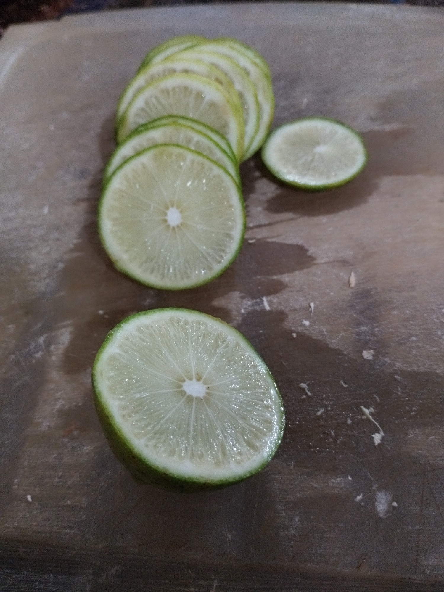 The image displays several freshly cut slices of lime on a wooden surface, likely a cutting board, in Lhokseumawe, Indonesia. The vibrant green rind and pale yellow-green flesh of the limes are clearly visible, with each slice showcasing its characteristic radial segments and a small white pith at the center.
A significant portion of the slices is arranged in an overlapping, slightly curved stack towards the upper left and center of the frame. Two additional, distinct circular slices are positioned in the foreground and mid-ground, separate from the main stack. The wooden board is a light brown, showing some natural grain and minor scuffs. Traces of moisture and small bits of lime pulp or residue are scattered across the surface, suggesting recent preparation. The lighting appears consistent with an indoor setting. There are no people or discernible text present in the image.