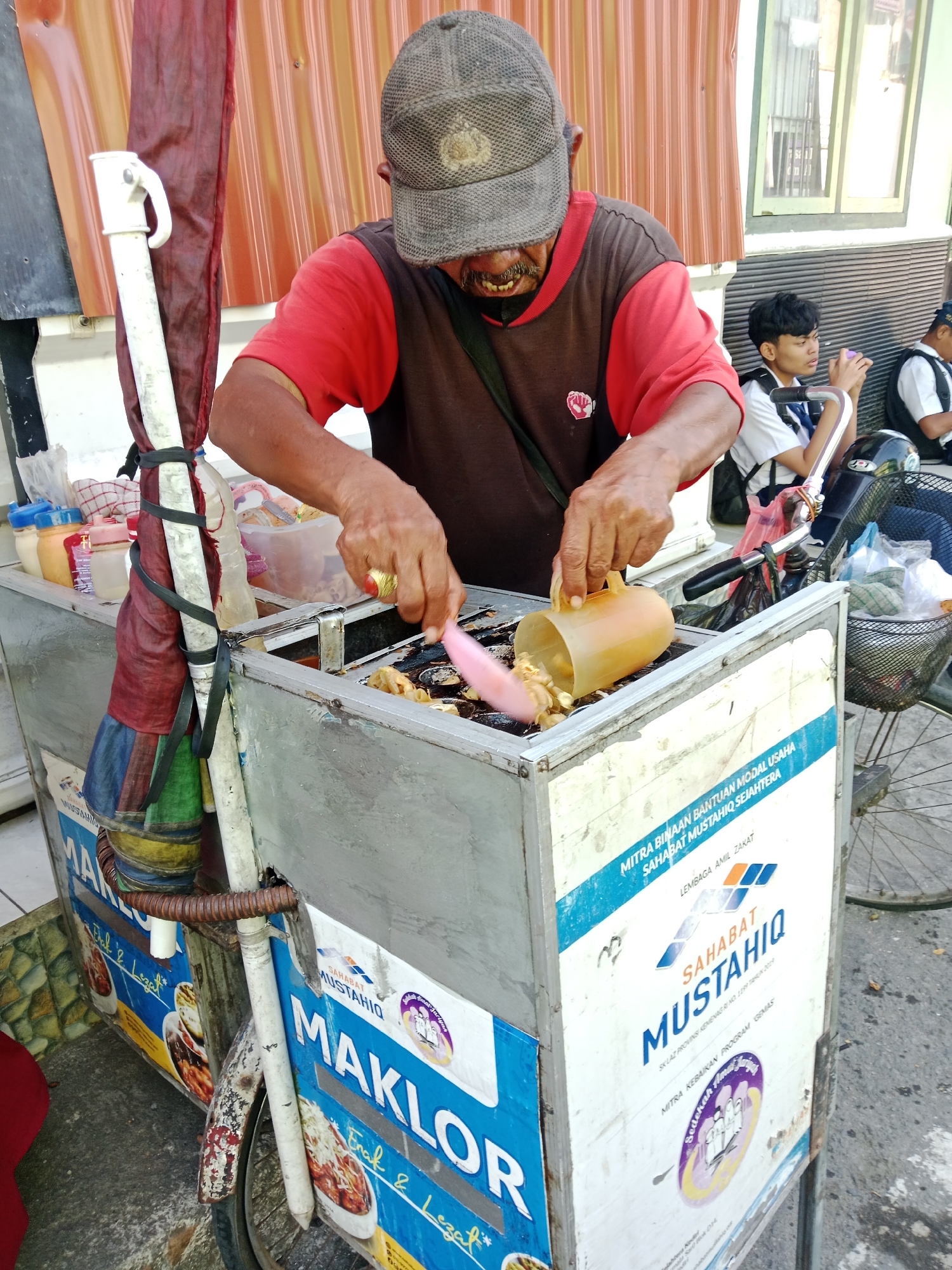 An elderly man wearing a red and brown shirt and a worn grey baseball cap is preparing food at a street cart in Kediri City, Indonesia. He is actively cooking, using a pink utensil to move food in a griddle and a yellow plastic pitcher to pour batter or oil. The cart is decorated with blue signage that includes text in Indonesian, featuring the words "MAKLOR," "Enak & Lezat," and "SAHABAT MUSTAHIQ." In the background, two young men in white shirts and backpacks are seated, one of them holding a purple object. The cart has a bicycle wheel attached to its side, suggesting it's mobile. The environment appears to be outdoors, with a building visible in the background.