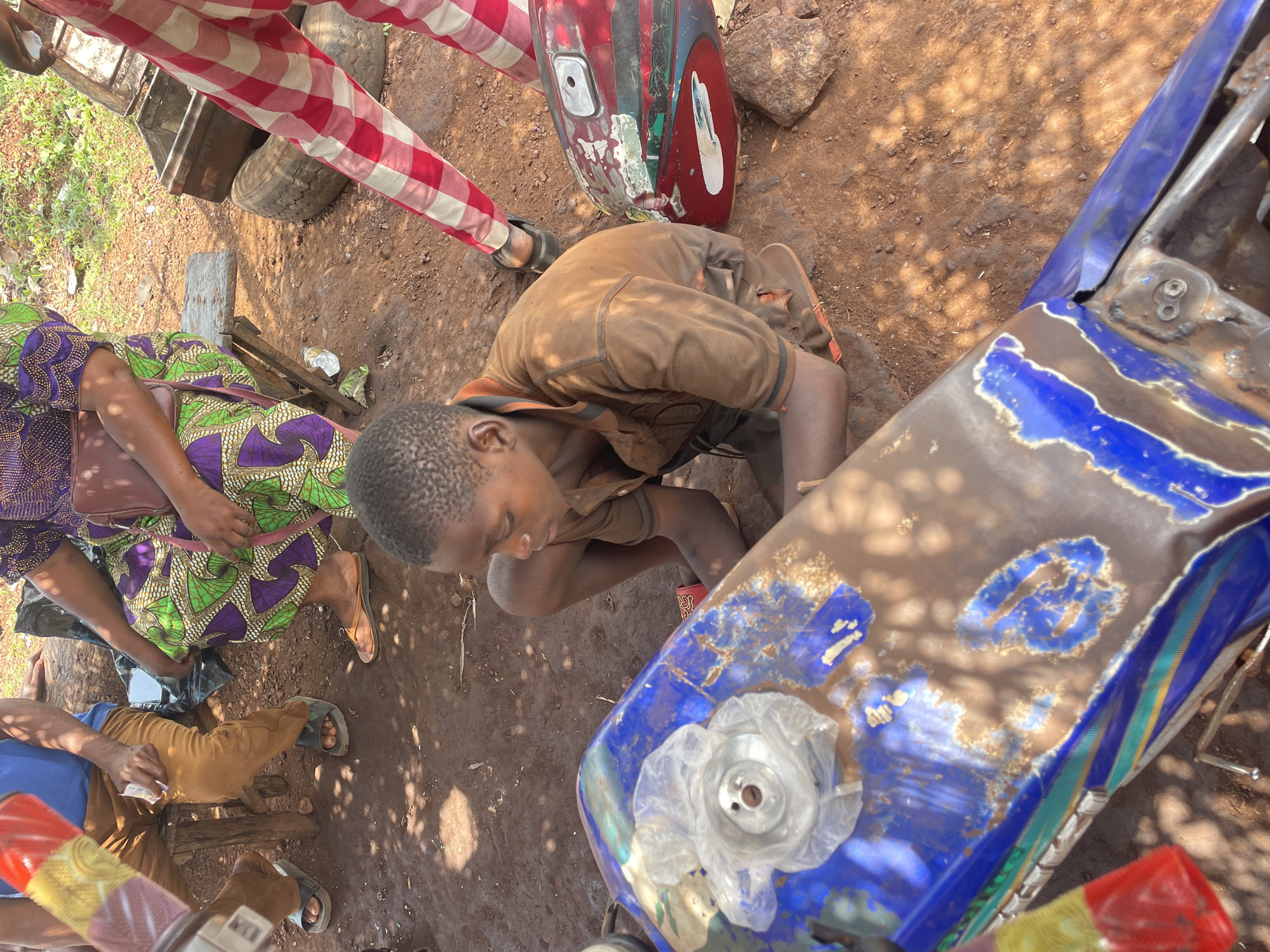 The image depicts an outdoor scene in Ogbe, Nigeria, under bright, direct sunlight, suggesting midday or early afternoon. The setting appears to be an informal workshop or gathering area, characterized by unpaved, reddish-brown dirt ground with scattered debris, rocks, and sparse vegetation.
At the center, a young boy or adolescent, wearing a brown short-sleeved shirt, is squatting or kneeling, intently focused on a heavily worn blue motorcycle fuel tank. The tank shows significant paint peeling, revealing the metal underneath, and a clear plastic bag covers the fuel cap opening. His posture suggests he is examining, repairing, or preparing the tank.
Several other individuals are partially visible around him. To the upper left, a person stands wearing red and white checkered pants. To the boy's left, a person dressed in a vibrant green and purple patterned skirt or dress is seated on a low wooden stool, holding a brown handbag. Further to the bottom left, another person in brown pants and a blue shirt is also seated on a low stool, holding what appears to be a small item or money. Feet and lower legs of an additional person are visible at the bottom right.
Behind the central boy, a red motorcycle fuel tank, also showing considerable wear, rests on the ground. The prominent, distinct shadows indicate clear, sunny weather. The overall scene conveys a casual yet focused activity, likely related to motorcycle maintenance or a roadside transaction, typical of a bustling informal outdoor environment. No visible text is discernible in the image.