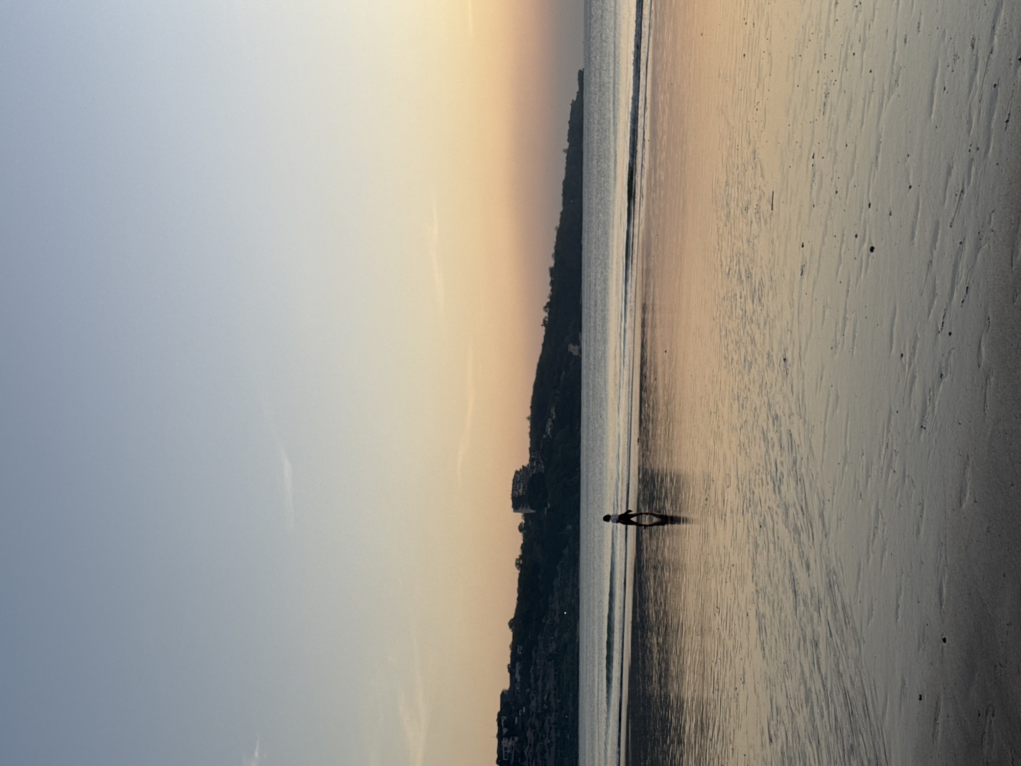 A solitary figure walks along a wet sandy beach reflecting the warm hues of a sunset over the ocean. The sky transitions from a pale blue at the top to a soft orange and yellow near the horizon. In the distance, a treed hillside is dotted with buildings, suggesting a coastal town. The water is calm, with small waves lapping the shore, and the wet sand creates a mirror-like surface. The scene captures a peaceful moment at dusk in Kuta Selatan, Indonesia.