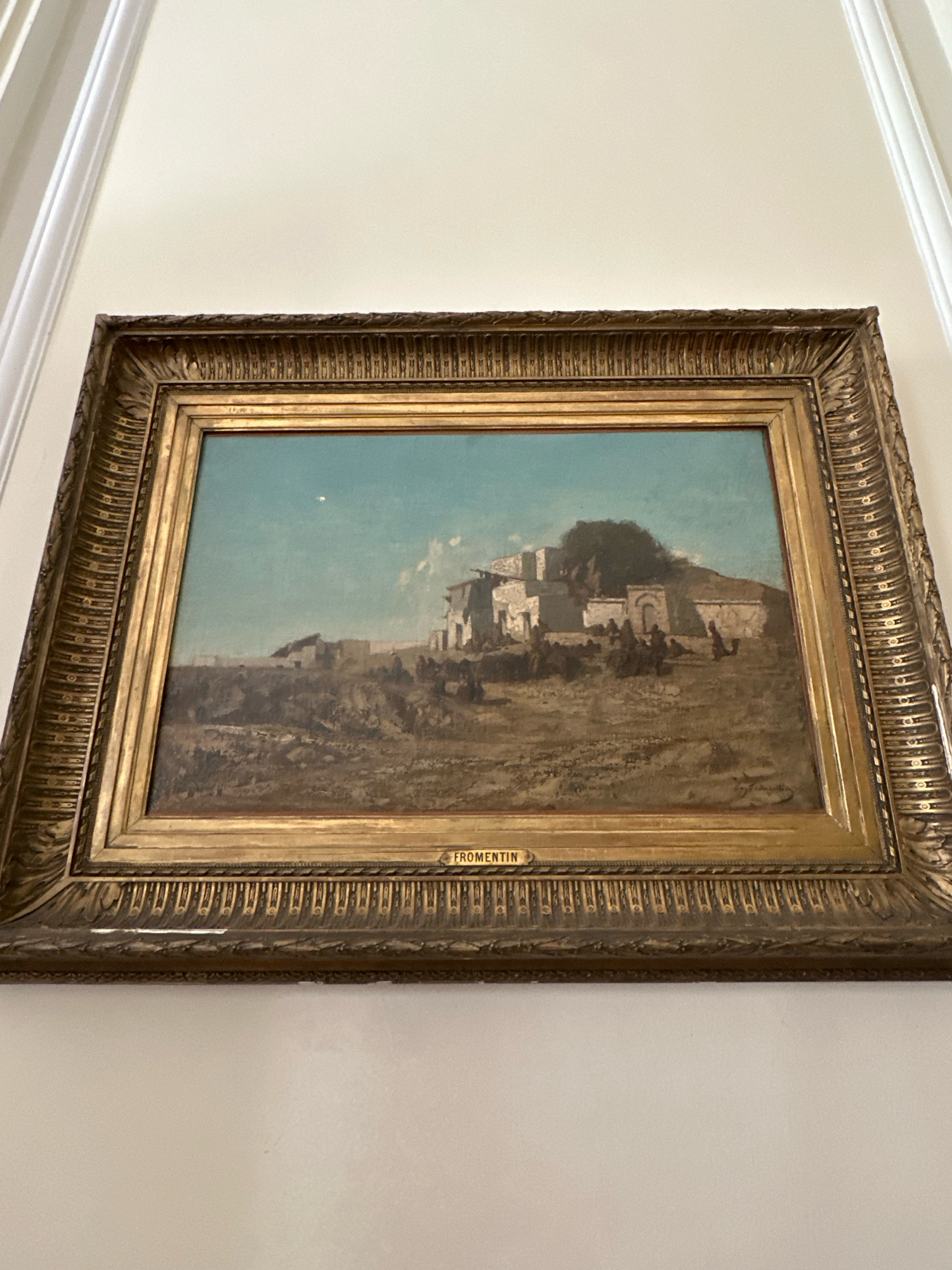 This is a photograph of a framed painting hanging on a plain, light-colored wall. The painting depicts an arid landscape with a village built on a hill. The village appears to be constructed of stone and has a somewhat dilapidated look. There are figures, possibly people, visible in the foreground and around the village, appearing as small, dark shapes. The sky is a light blue with some wispy clouds. The painting is signed "FROMENTIN" on a small plaque attached to the frame. The frame itself is ornate and appears to be gilded. The painting is displayed at an angle, and the wall has white molding at the top. There is no indication of the painting's location in the city of Belgrade, Serbia.