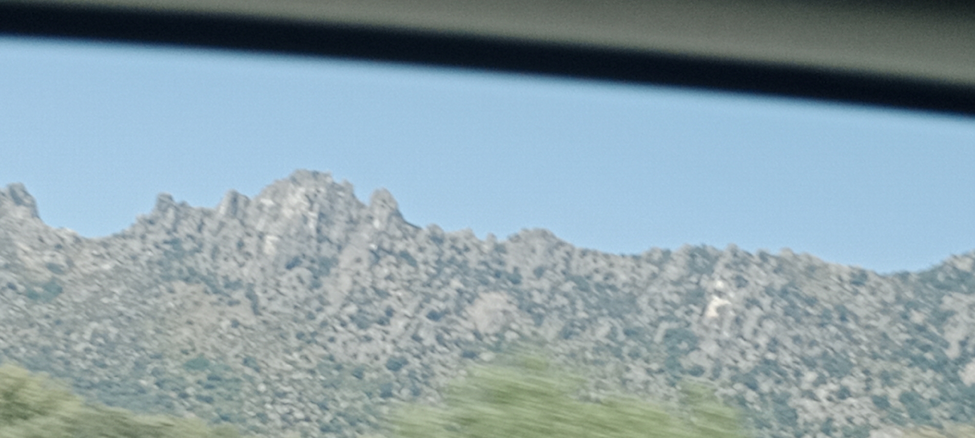 This is a horizontal, medium-shot, landscape photograph of the mountains near La Cabrera, Spain, under a clear blue sky. The image is slightly blurred, suggesting it was taken from a moving vehicle. The foreground is dominated by the rugged, rocky texture of the mountain range, which appears sparsely vegetated with small shrubs. The peaks of the mountains are jagged and irregular, forming a distinct silhouette against the sky. The lower portion of the image shows a blurred expanse of green, likely trees or low-lying vegetation. A dark, horizontal band at the top of the image suggests the view is framed by the interior of a vehicle, possibly the car's roofline or a sun visor.