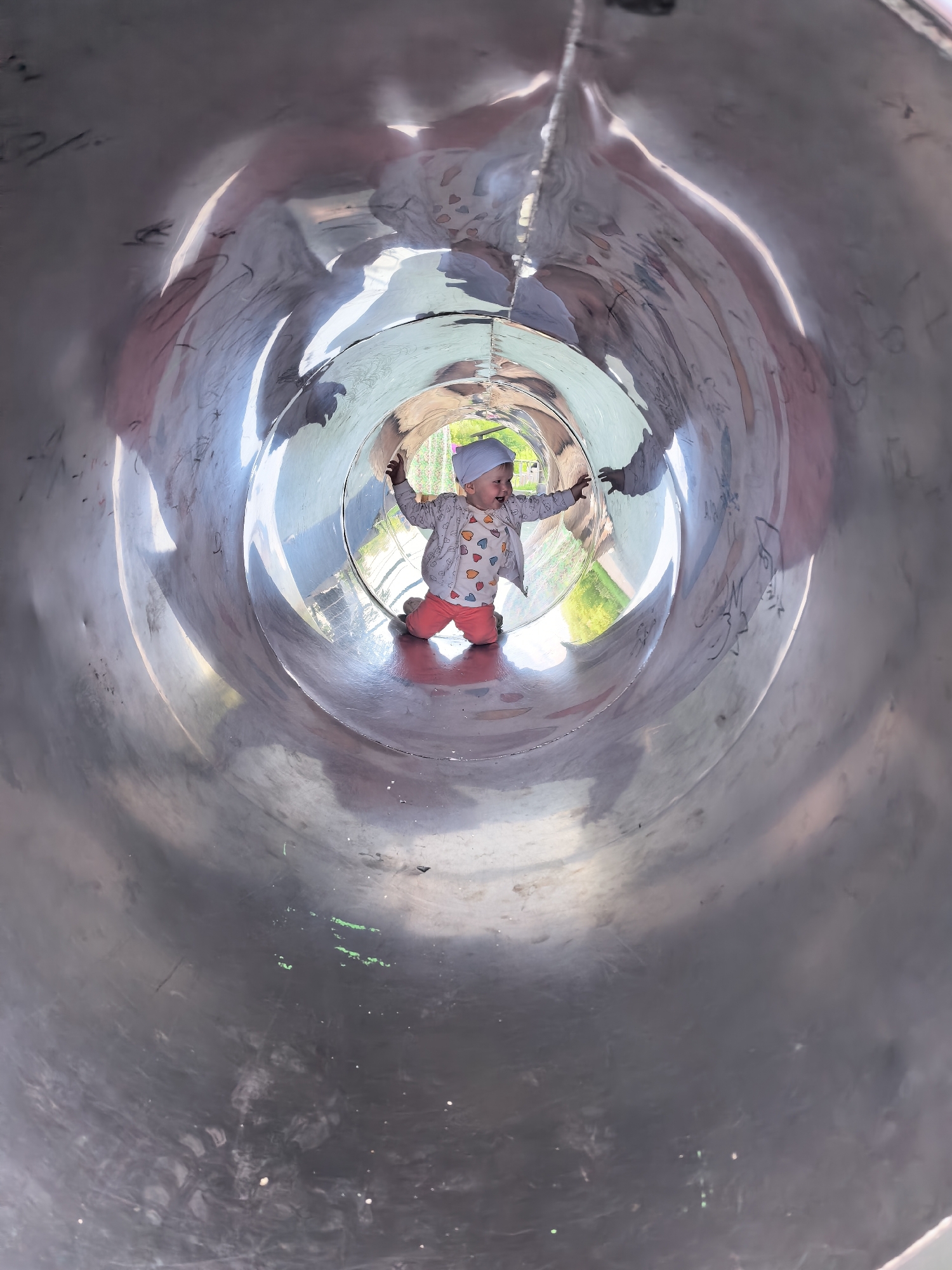 A toddler is kneeling inside a metal tube slide at a playground. The toddler is wearing a light blue hat, a long-sleeved shirt with colorful heart patterns, and orange pants. The toddler is looking to the left and has their arms outstretched. The inside of the slide is decorated with drawn pictures and has a reflective surface, showing distorted reflections of the surroundings and the child. Sunlight streams in from the opening of the slide at the far end, illuminating the child and the interior. The exterior of the slide appears to be made of polished metal. This image was taken at a playground in Sterlitamak, Russia.