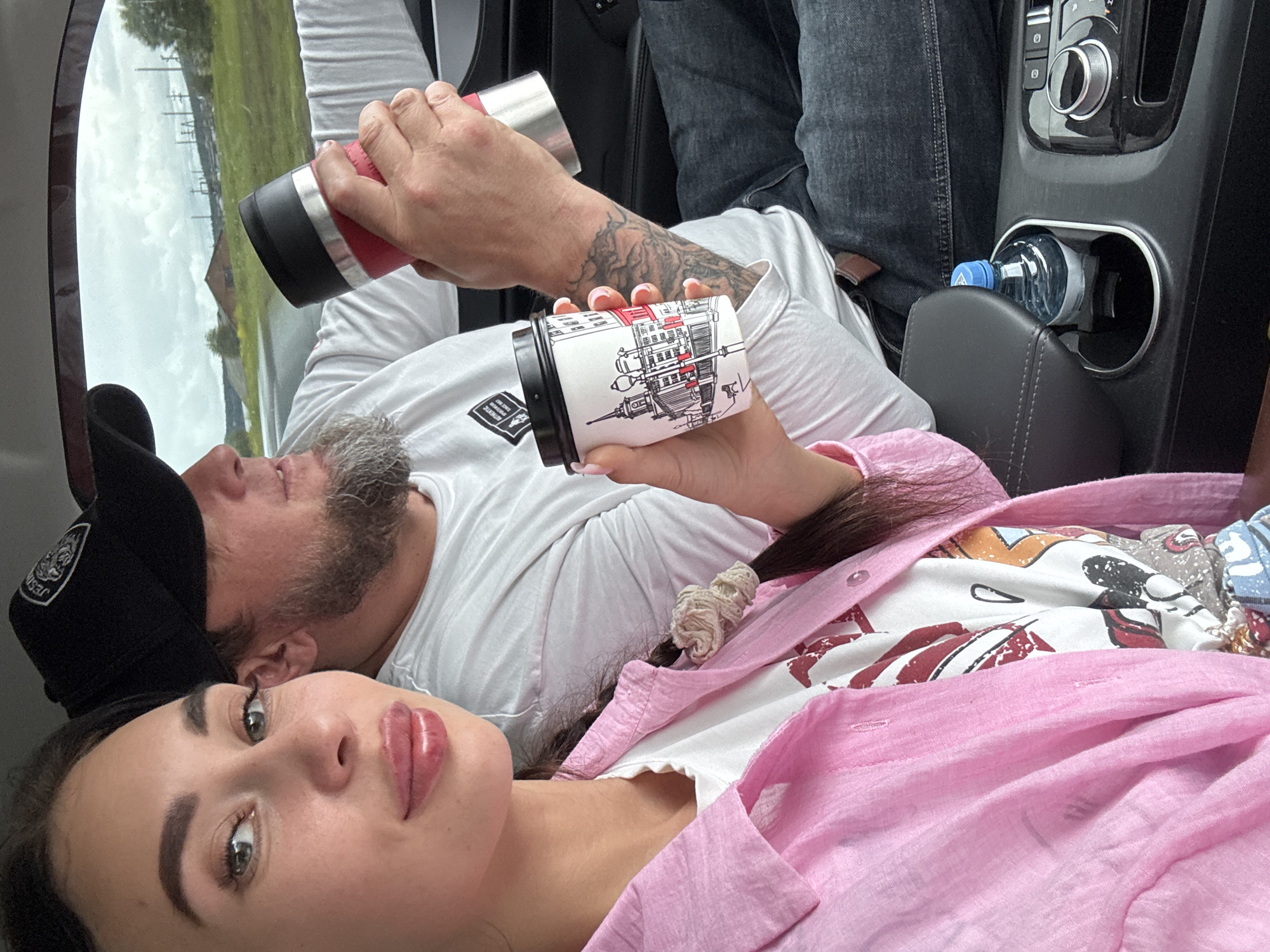 A woman and a man are in a car. The woman is in the foreground, looking at the camera, while the man is in the background, looking forward. The woman is wearing a pink shirt and a white t-shirt. She has long dark hair and is holding a coffee cup. The man is wearing a black baseball cap and a white t-shirt. He has a beard and is holding a red thermos. The car interior is visible, including the dashboard and a cup holder with a bottle of water. The background outside the car shows a grassy landscape with some industrial structures and a cloudy sky. The image appears to have been taken in Pervomaiskoe, Russia.
