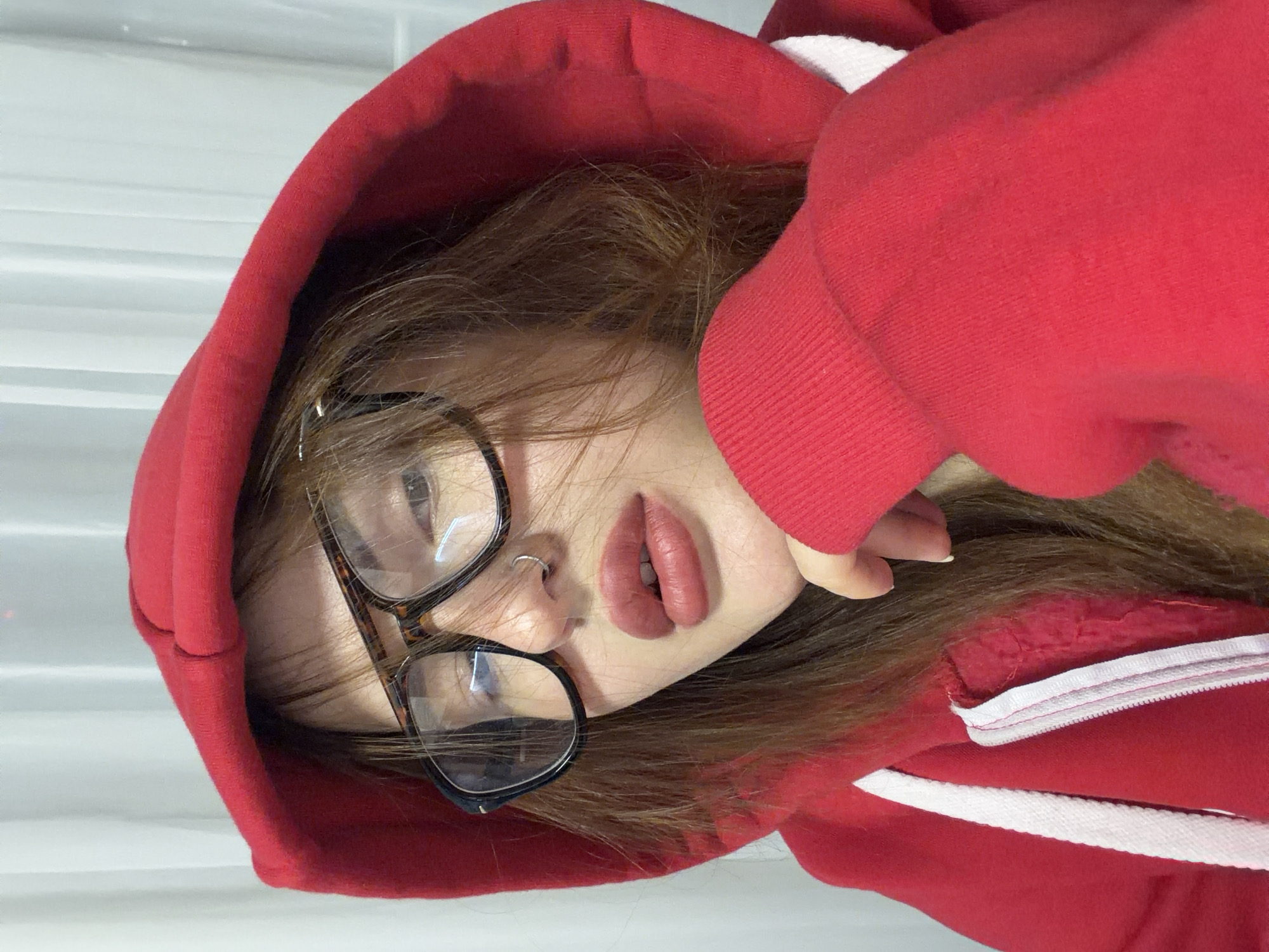 A close-up, rotated portrait features a person, likely female, with long, light brown hair, looking directly at the camera. They wear a vivid red hooded sweatshirt, featuring white drawstrings and internal lining accents. The person has a silver hoop nose piercing and is wearing eyeglasses with distinctive tortoise-shell frames. Their lips are painted a muted pinkish-red and are slightly parted, revealing just the top row of teeth, which conveys a pensive or alluring expression. Part of a hand is visible near the back of their head, likely their own. The background consists of a plain, light-colored, possibly white, surface with subtle horizontal lines, indicating an indoor setting. No text is visible in the image. This image is from Krasnodar, Russia.