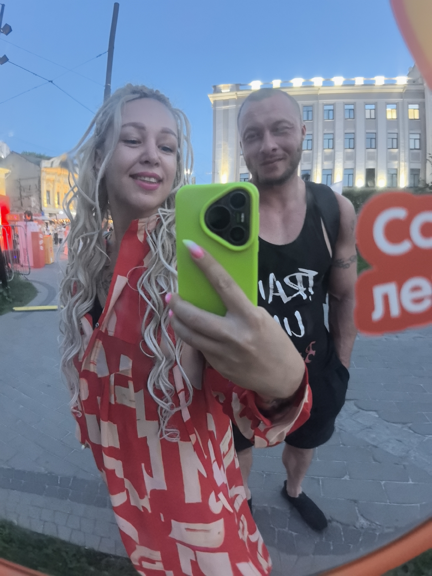 The image shows a mirrored reflection of a couple in an outdoor urban setting. In the foreground, a woman with blonde dreadlocks is holding up a bright green smartphone, taking a selfie. She is wearing a red and beige patterned shirt. To her right, a man with short hair and a beard is visible, wearing a black tank top. Behind them, a large building with many windows and bright lights can be seen. The ground appears to be paved. The reflection also shows a red sign with white lettering that is partially visible. The overall impression is a candid moment captured at dusk in a public space. The building in the background is the Bolshaya Pokrovskaya Street tram depot, a historical landmark in Nizhny Novgorod, Russia.