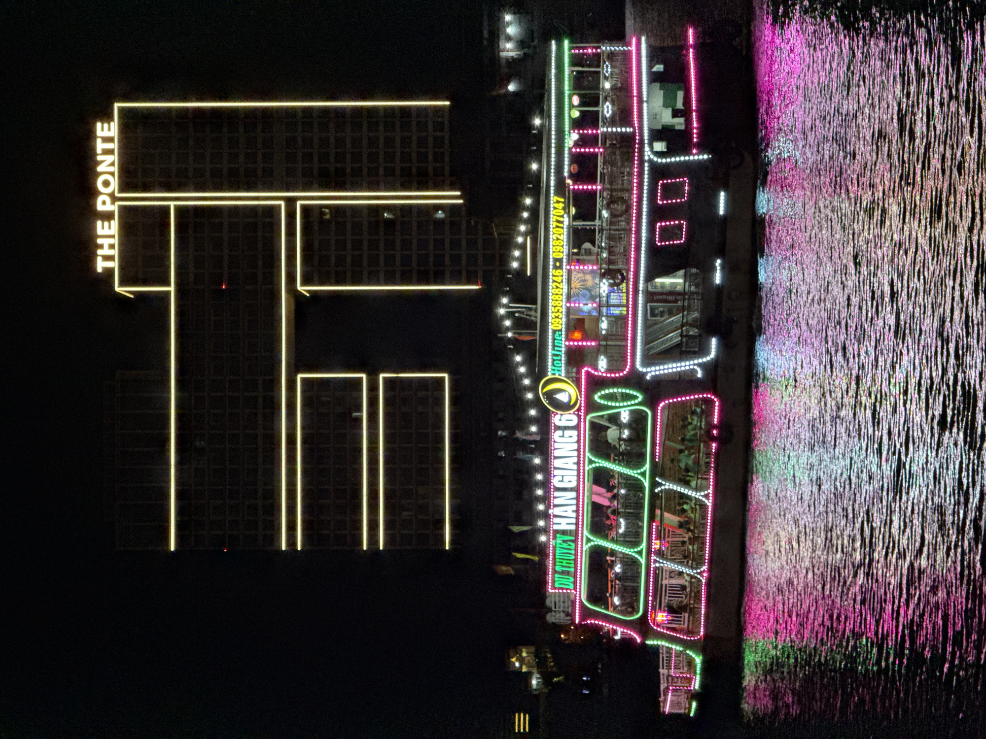 The image captures a vibrant night scene along a riverfront in what appears to be a bustling city, confirmed as Đà Nẵng, Vietnam. On the left, a tall, dark building is outlined by striking yellow neon lights, with the name "THE PONTE" displayed vertically in matching yellow lettering.
In the center of the frame, a multi-deck riverboat or cruise ship is brightly illuminated with an array of colorful LED lights in pink, green, white, and yellow, giving it a festive appearance. People can be seen as silhouettes on the boat's various decks, suggesting an evening activity such as dining or sightseeing. The boat features extensive lighting patterns and visible seating areas.
The name "DU THUYEN HAN GIANG 6" is prominently displayed on the side of the boat in green and yellow text, along with "Hotline: 0935888246 - 0982077047" in yellow.
To the right, the dark water of the river reflects the myriad of lights from the boat and possibly the distant city, creating shimmering patterns of pink, green, and white across its surface. The clear night sky and the distinct reflections indicate good weather. The overall atmosphere is lively and inviting, typical of a nighttime entertainment or tourist experience in an urban setting.
